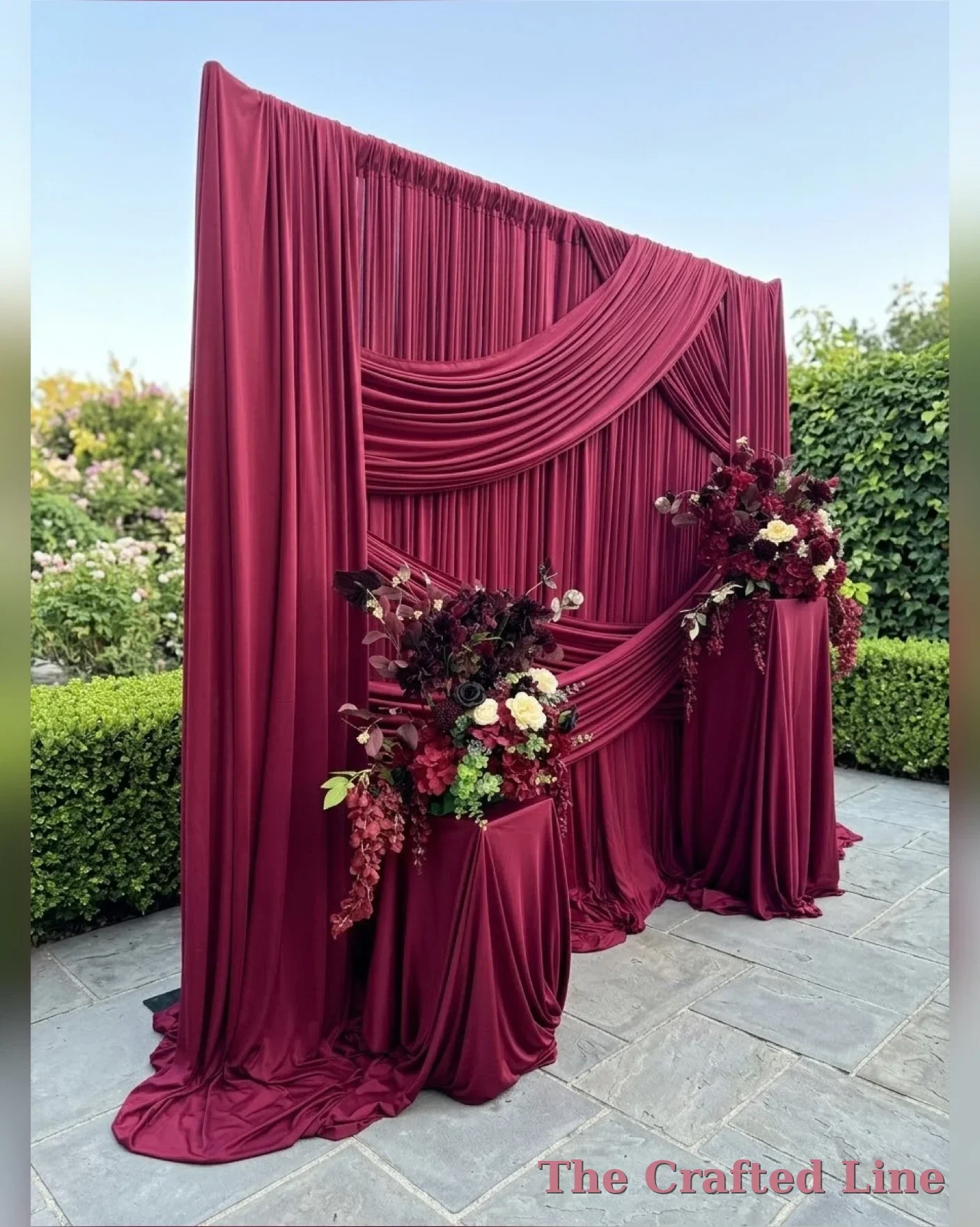 A decorative red fabric backdrop with draped panels and floral arrangements at the base, set outdoors on a patio with bushes and trees in the background.