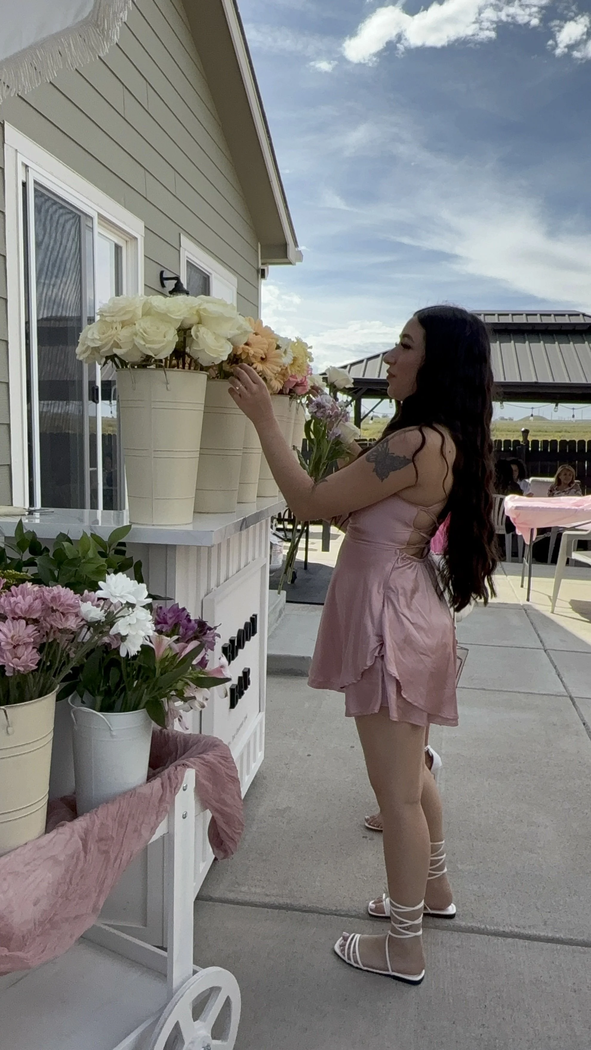 A woman with long dark hair, wearing a pink dress and white sandals, arranging flowers on a white table outdoors in a backyard or patio area. There are flower pots with large white, pink, and peach flowers. The sky is partly cloudy, and there are oth