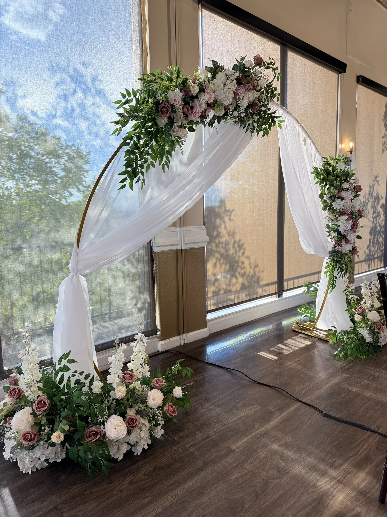 Wedding arch decorated with white and pink flowers and greenery, draped with white fabric indoors