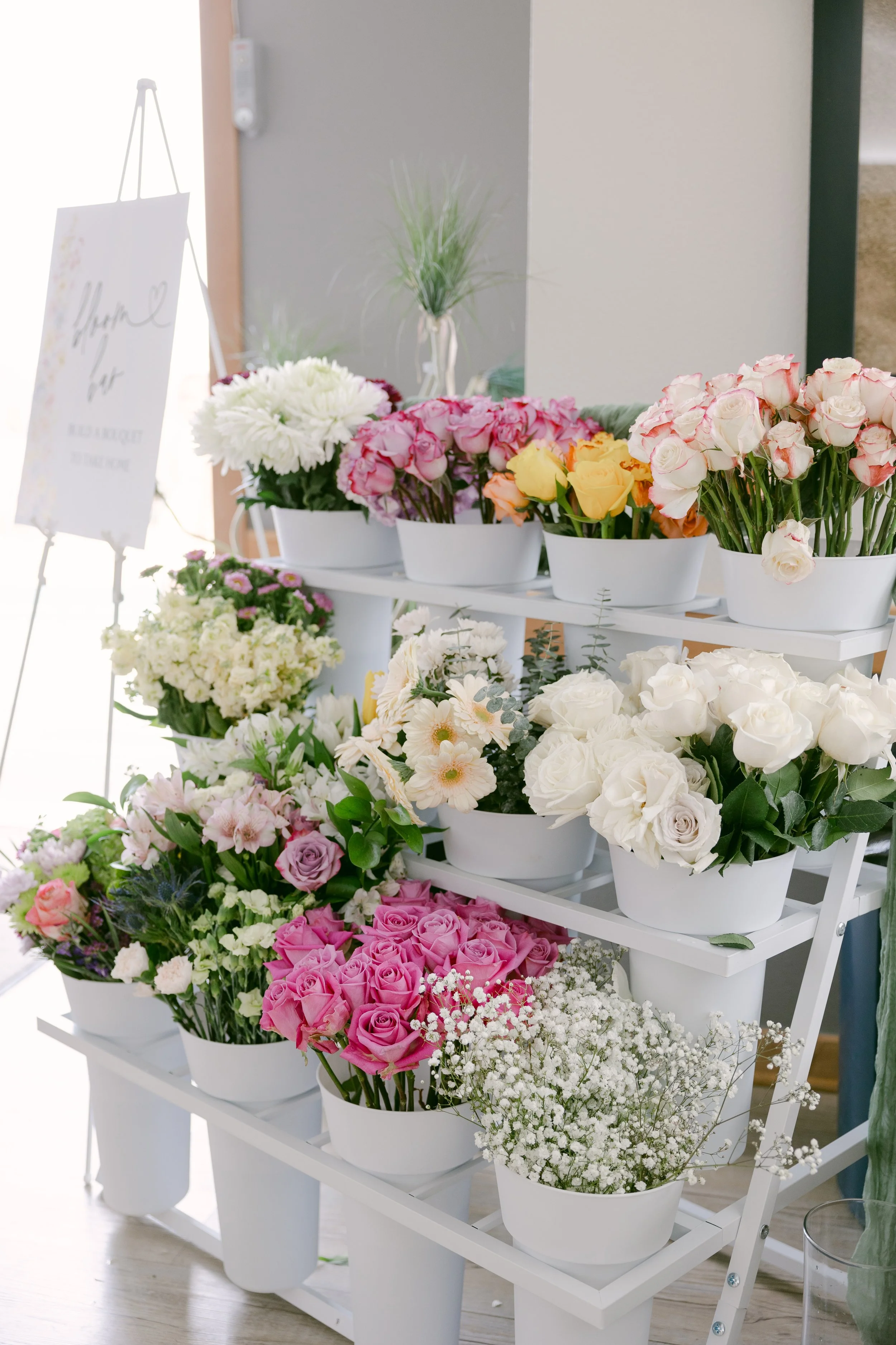 White shelf with white flower pots filled with various colorful flowers, including roses, baby's breath, and other blooms, in a bright room.