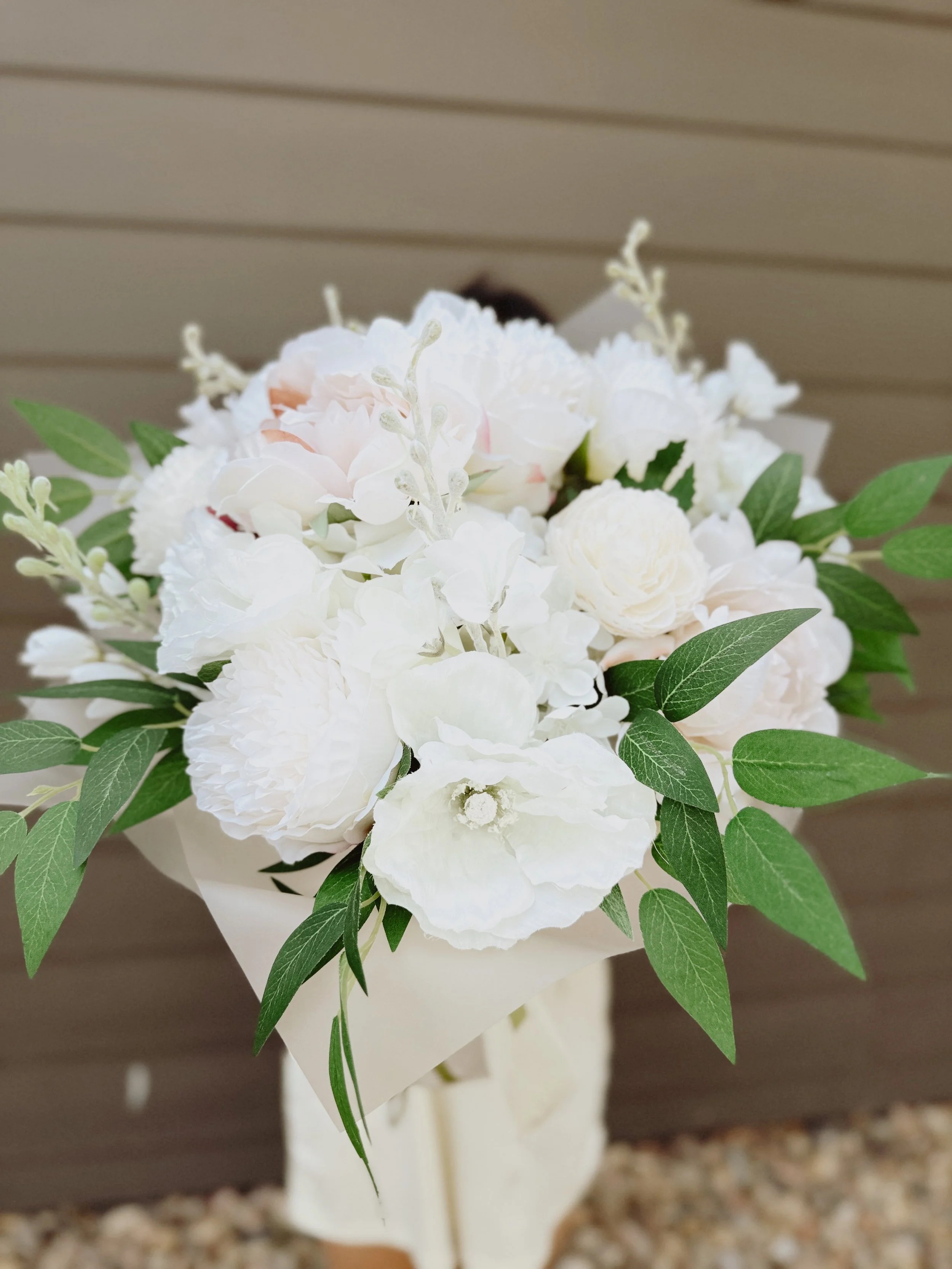 A bouquet of white flowers with green leaves, wrapped in white paper, against a gray wall background.