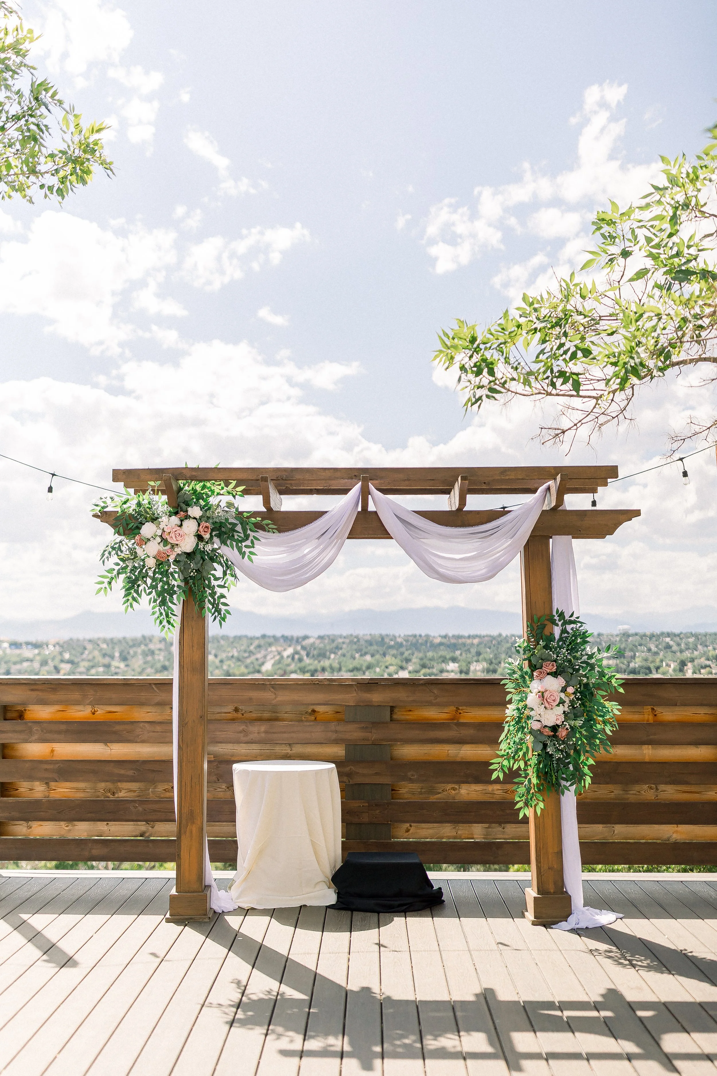 Outdoor wedding arch on a wooden deck with floral decorations and draped fabric, set against a scenic sky with clouds and trees.