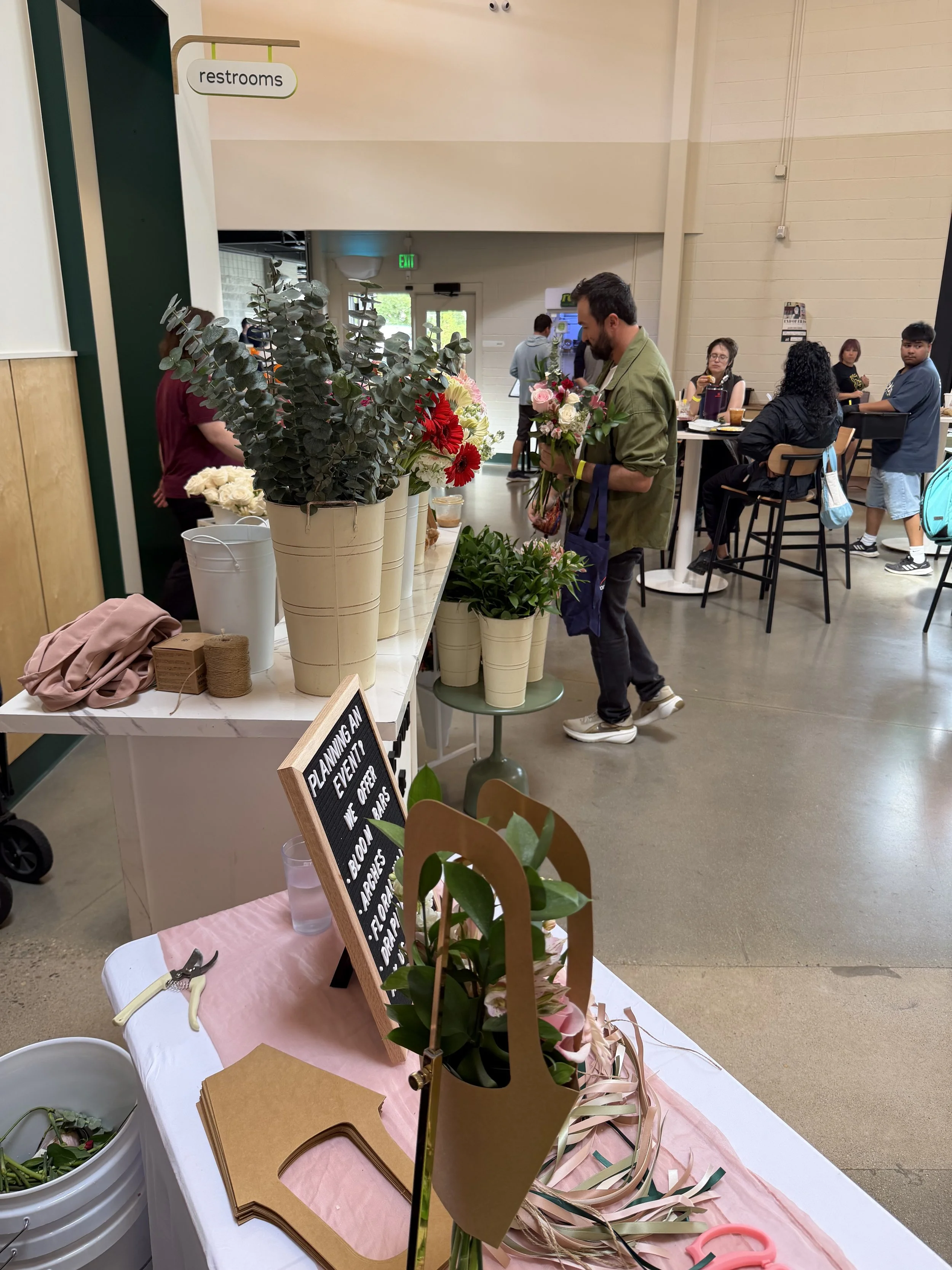 People shopping for flowers at an indoor market, with bouquets and floral supplies displayed on tables.