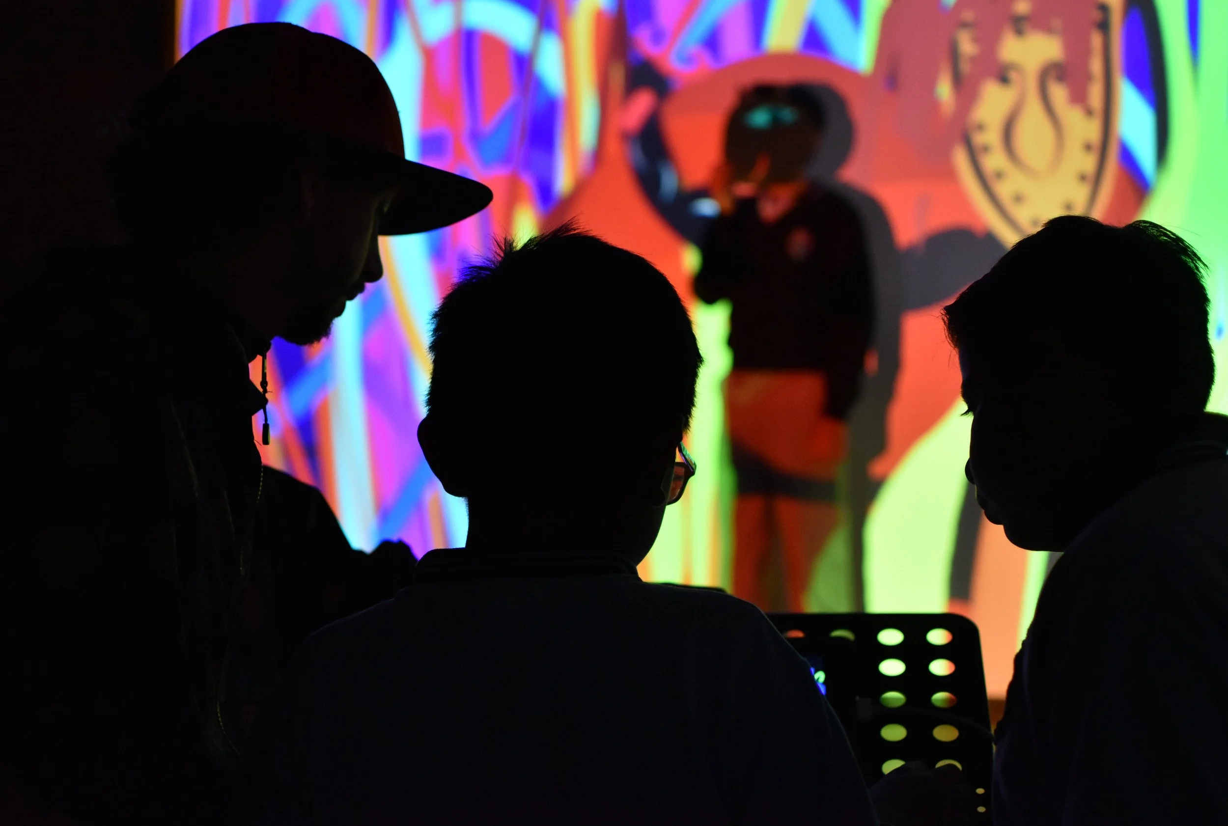 Silhouettes de varias personas en un entorno oscuro con pantallas y luces de colores, probablemente en un evento o presentación tecnológica.