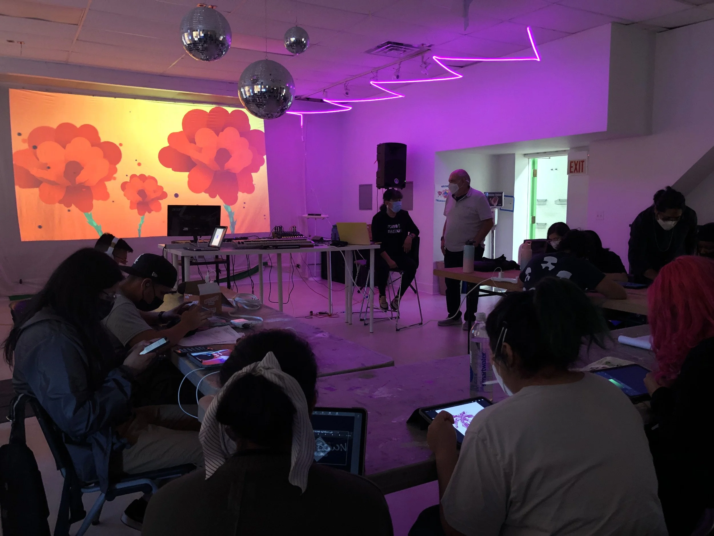 Una sala con muchas personas usando dispositivos electrónicos, en la pared hay una proyección con flores de color naranja y fondo amarillo, y varias bolas de discoteca colgadas del techo, con luces neón rosadas.