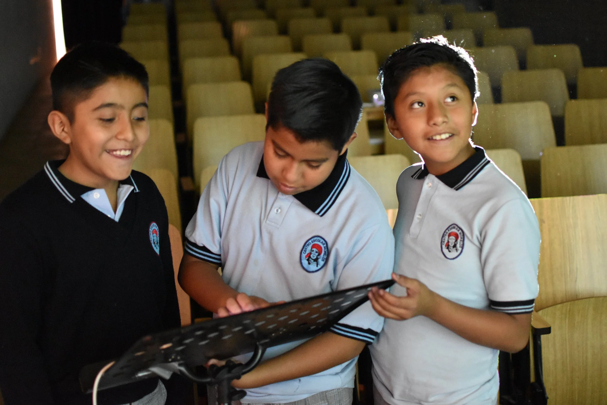 Tres niños con uniforme escolar observando algo en un dispositivo electrónico en un aula con sillas vacías de fondo.