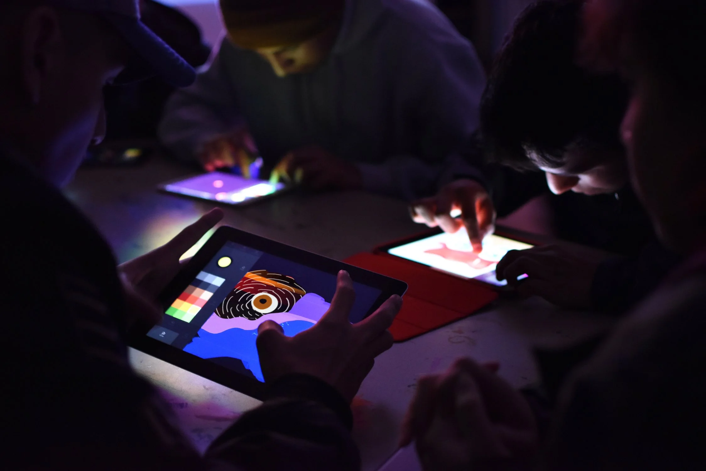 Personas usando tablet y teléfonos en un ambiente oscuro, visualizando arte digital y colores vibrantes.