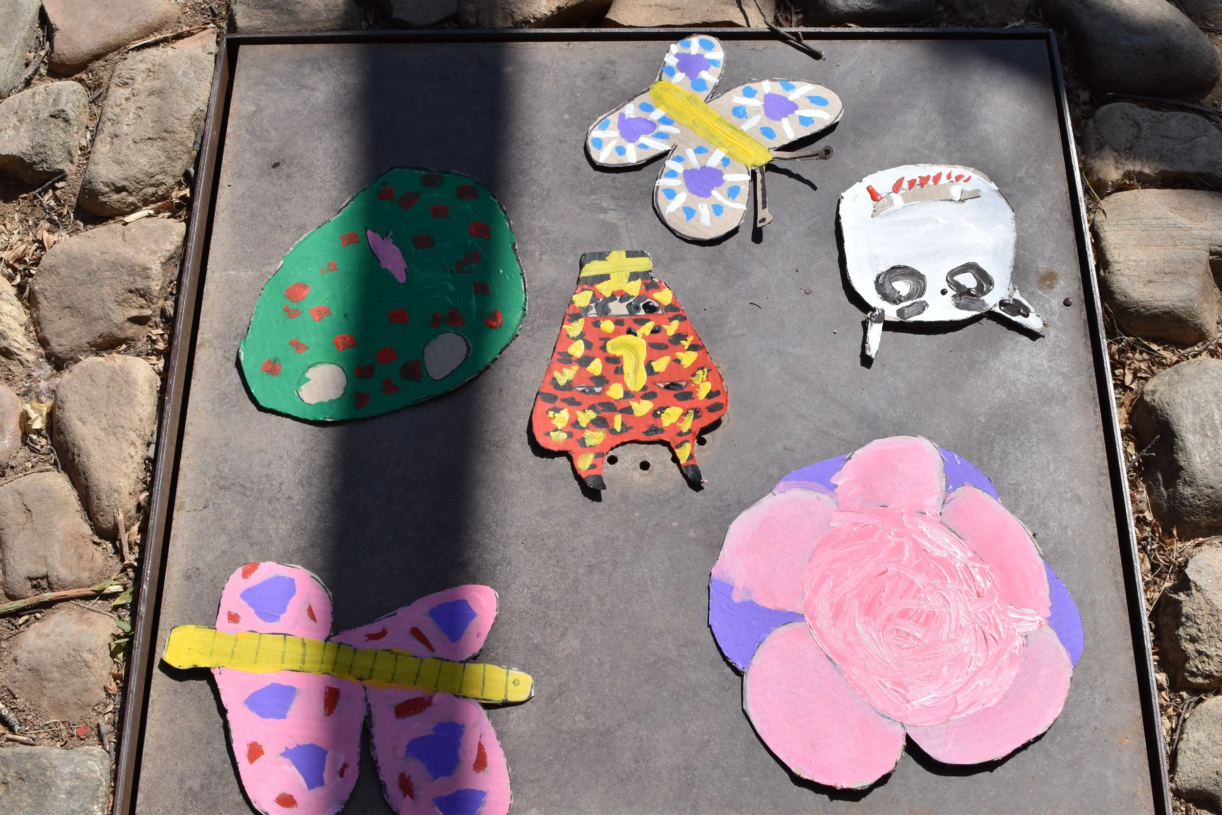 Cuatro mariposas de cartón pintadas con colores brillantes y patrones diversos, colocadas en una superficie de piedra rodeada de piedras.