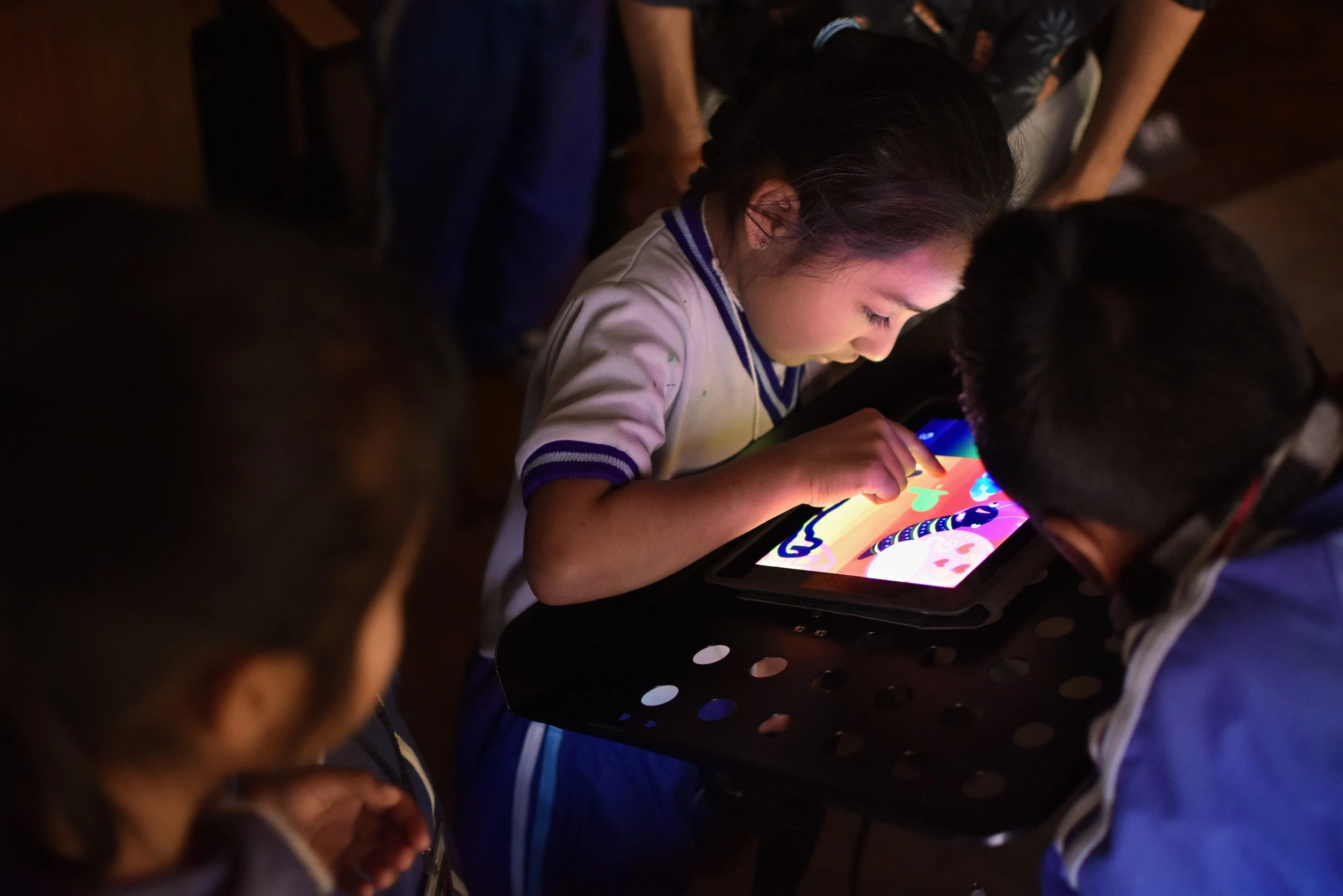 Niña usando una tableta en un ambiente oscuro, rodeada por otras personas.