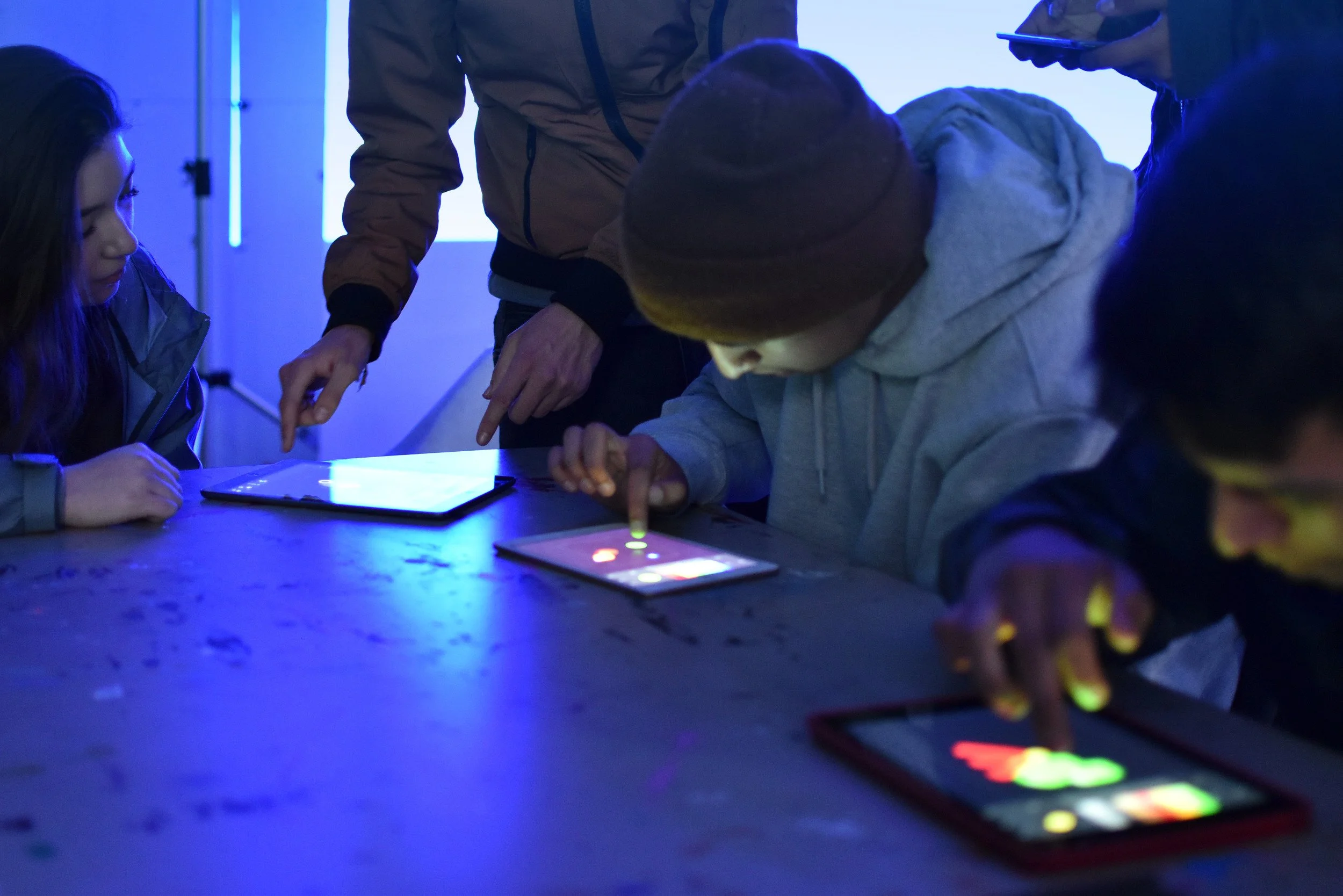 Grupo de personas en una mesa, usando dispositivos electrónicos en un entorno con iluminación azul y pantalla brillante.