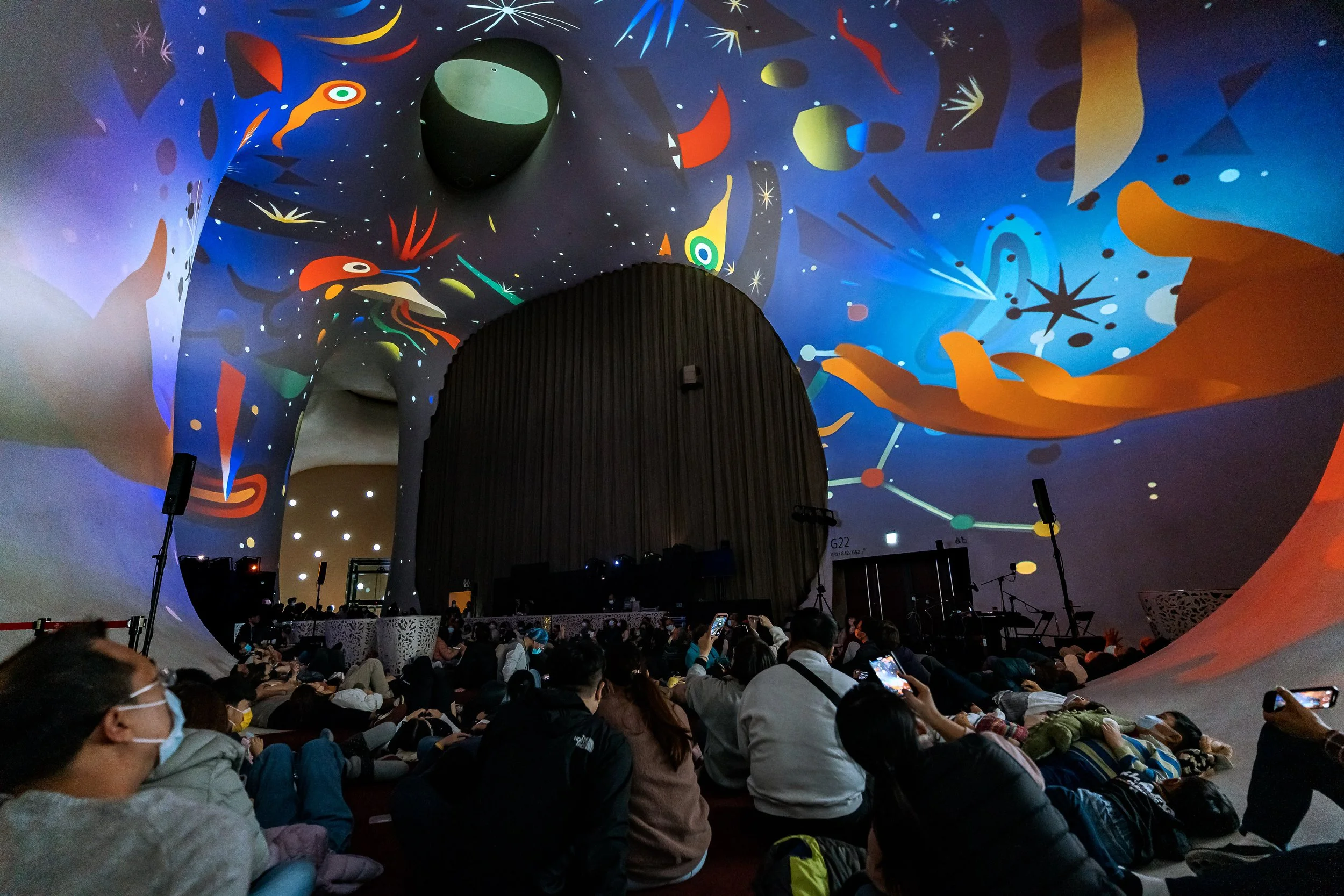 Interior de un auditorio con un techo y paredes decorados con arte espacial y estrellas, lleno de personas sentadas y algunas tomando fotos, algunas llevan mascarillas.