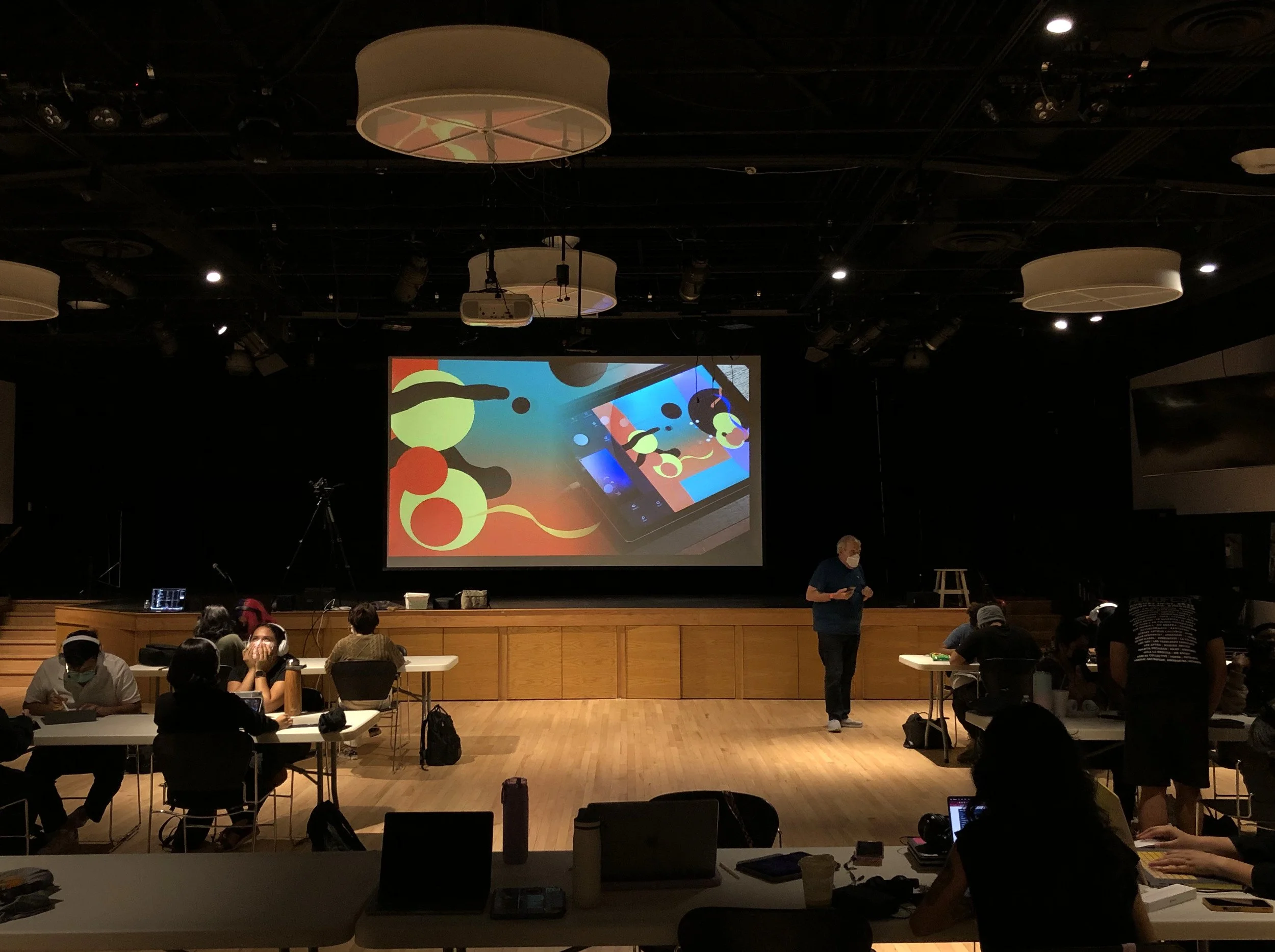 Escenario con proyector y pantalla en un auditorio, personas sentadas en mesas con laptops y audífonos, varias personas usando mascarillas, techo oscuro con luces y altavoces.