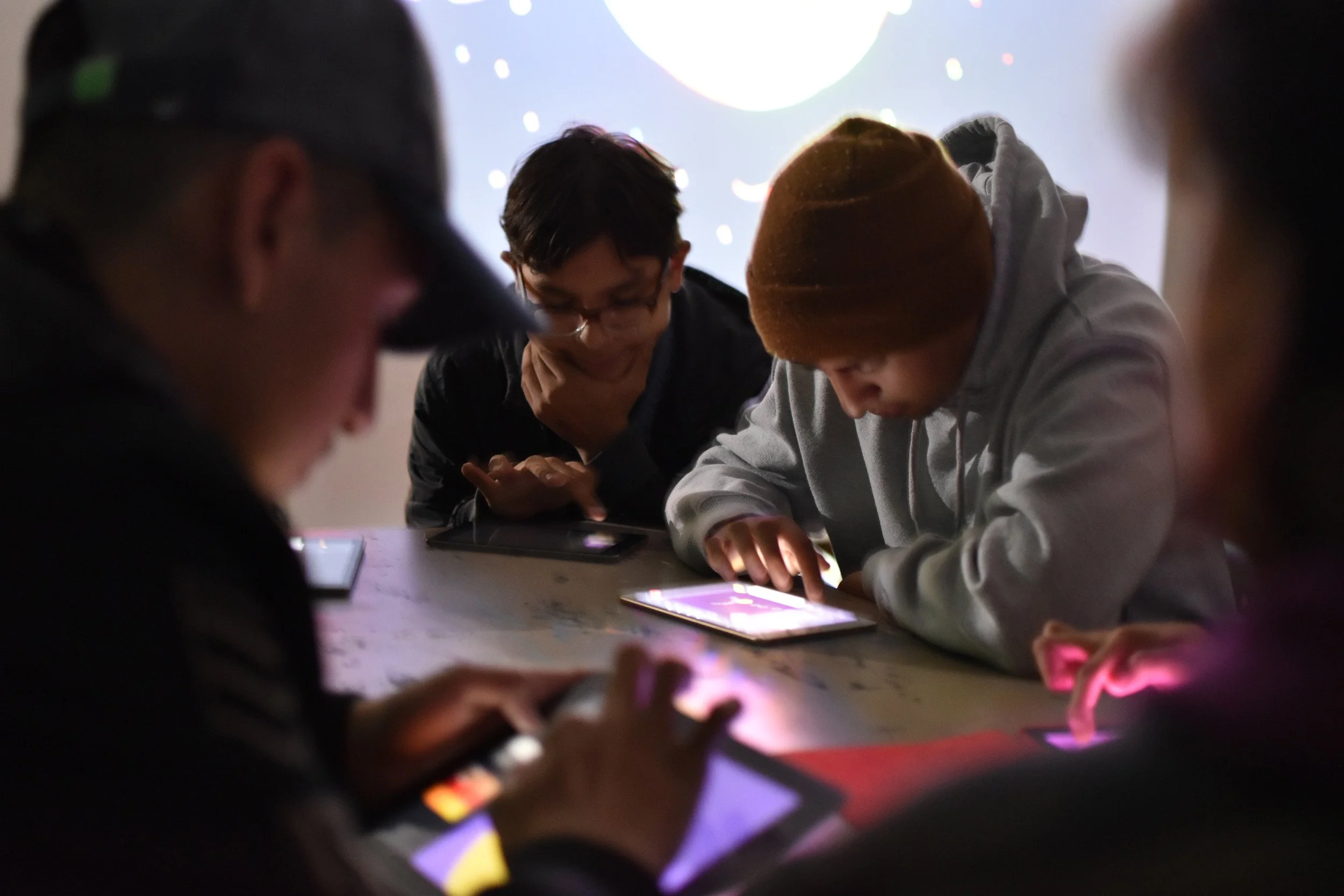 Grupo de jóvenes sentados en una mesa en un entorno oscuro, usando dispositivos electrónicos como teléfonos y tablets, con un fondo con una pantalla brillante.