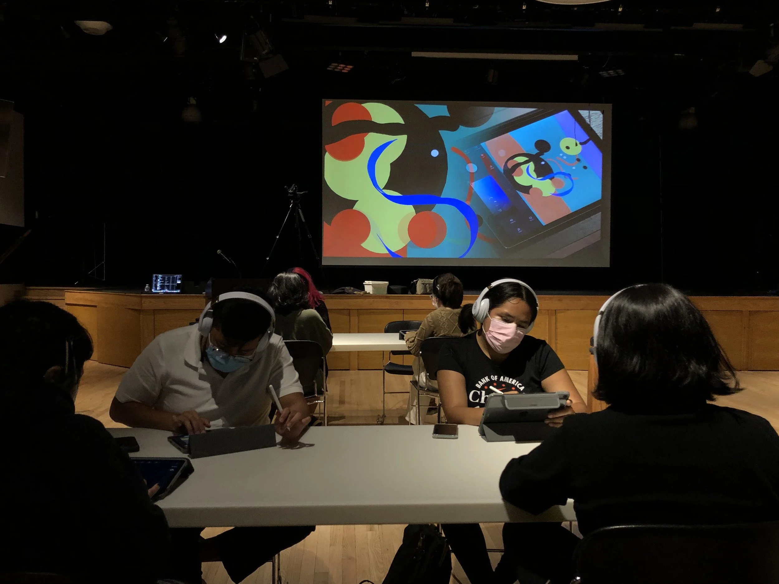 Una sala con varias personas usando dispositivos electrónicos, una pantalla grande muestra ilustraciones coloridas de tecnología y dispositivos móviles.