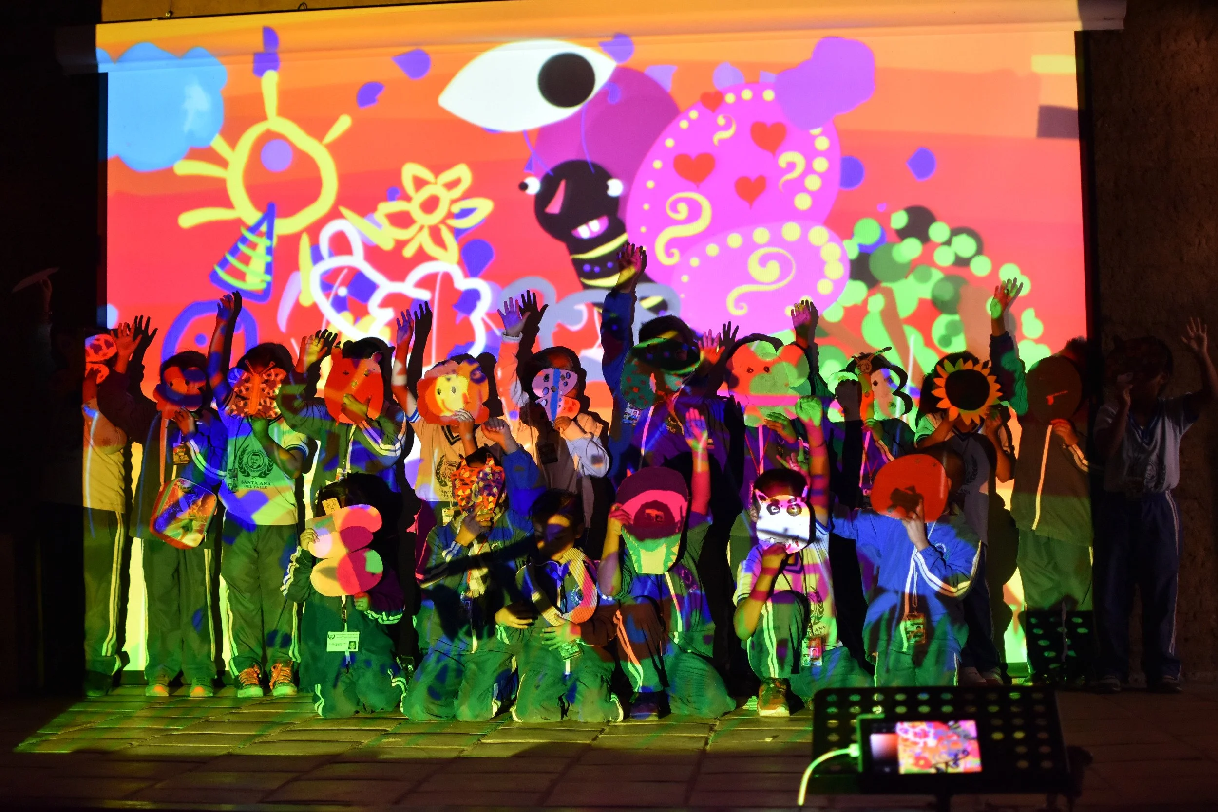 Niños y adultos en un escenario con proyección colorida y dibujos de animales y corazones, participando en una actividad artística.