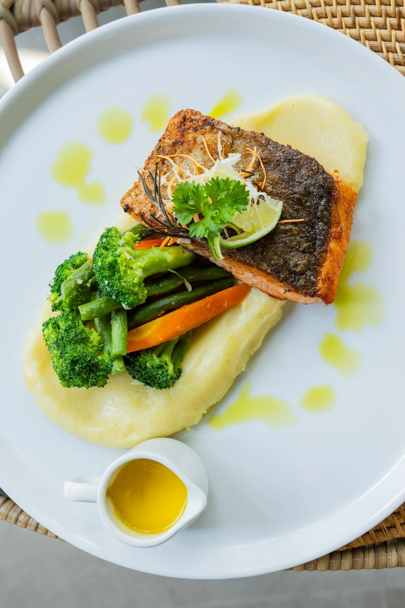 A plate of cooked salmon with crispy skin, served with mashed potatoes, cooked broccoli, and sliced carrots, garnished with lemon, parsley, and shredded cheese, with a small pitcher of yellow sauce on the side.