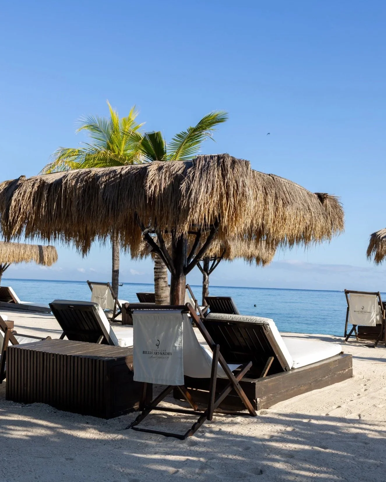 Beach scene with thatched umbrellas, lounge chairs, and a calm ocean under a clear blue sky.