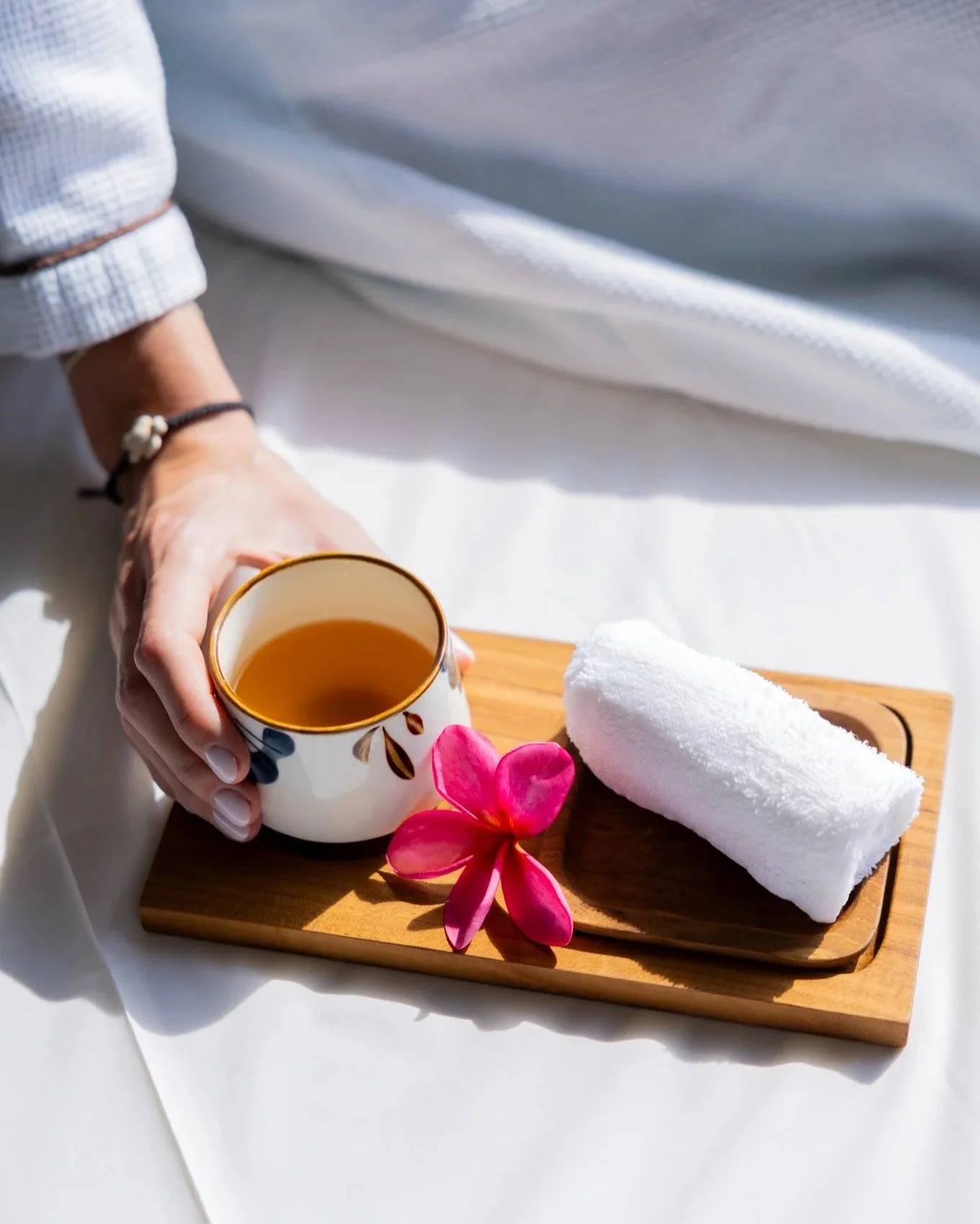 A person holding a ceramic cup of tea, with a rolled white towel and a pink flower on a wooden tray, set on white fabric.
