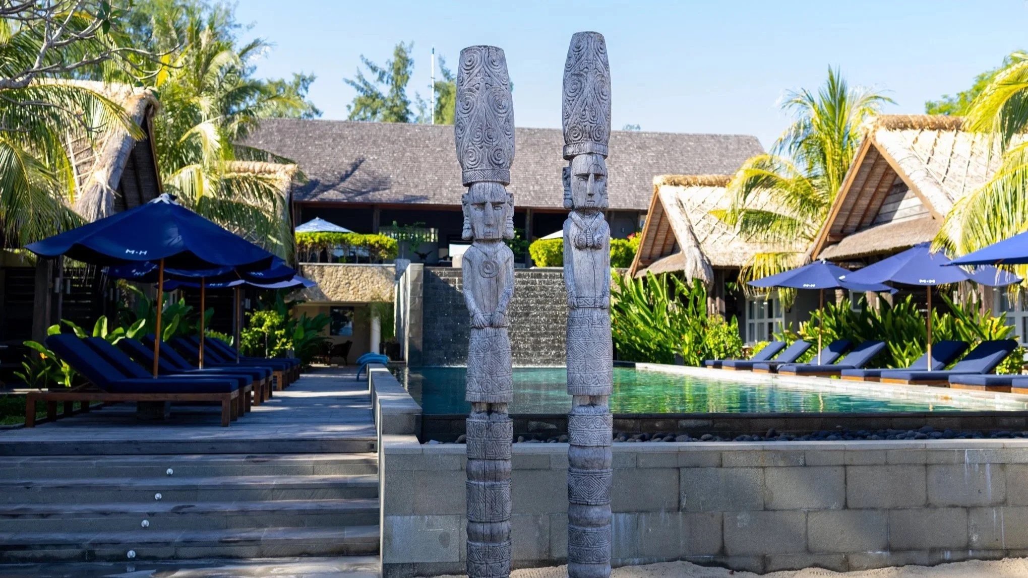 A tropical resort pool area with wooden statues in the foreground, surrounded by palm trees, lounge chairs with blue umbrellas, and thatched roof buildings in the background.