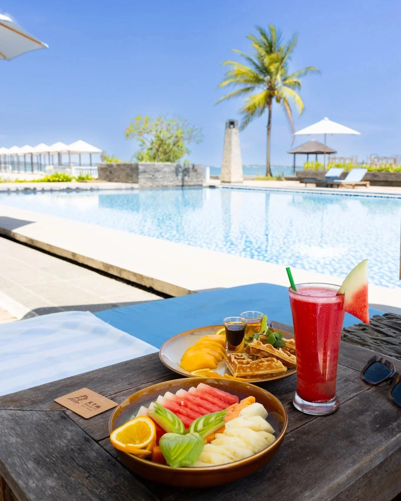 A tropical poolside scene with a table of breakfast foods including a juice, fresh fruit, waffles, and syrup, with pool, palm trees, umbrellas, and beach in the background.