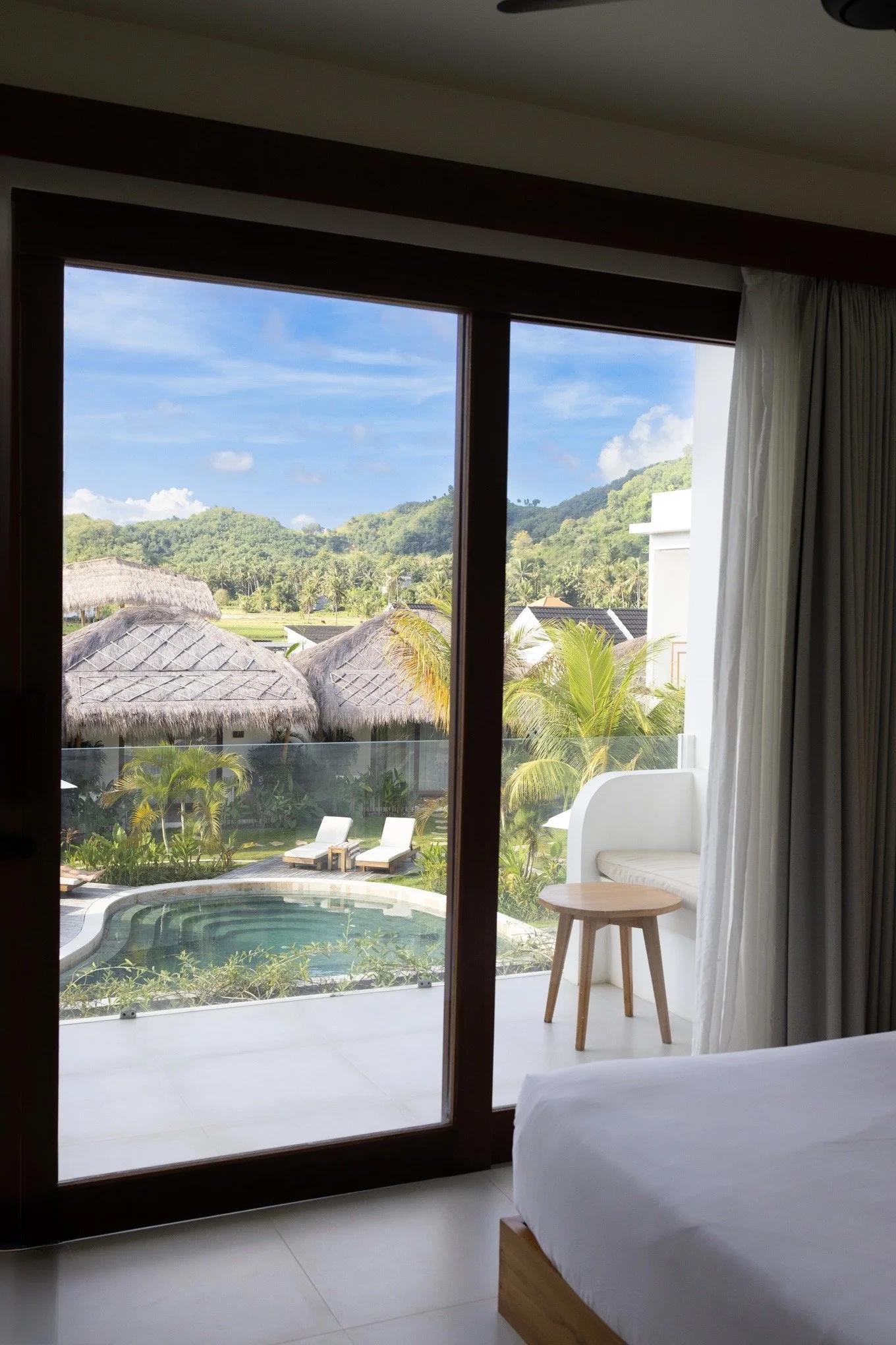 A view from inside a bedroom looking out through sliding glass doors. Outside, there is a pool with lounge chairs, lush green trees, thatched-roof huts, and hills in the distance under a partly cloudy sky.