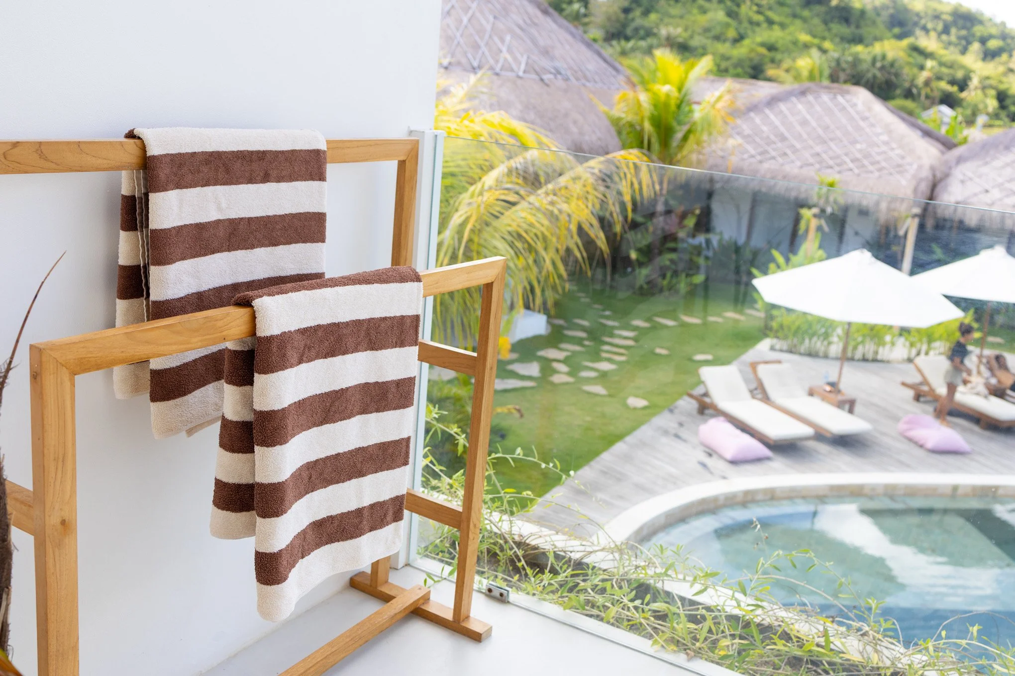 Bathroom with striped towel rack and towel, overlooking a balcony with view of tropical plants, roofed structures, and pool area.