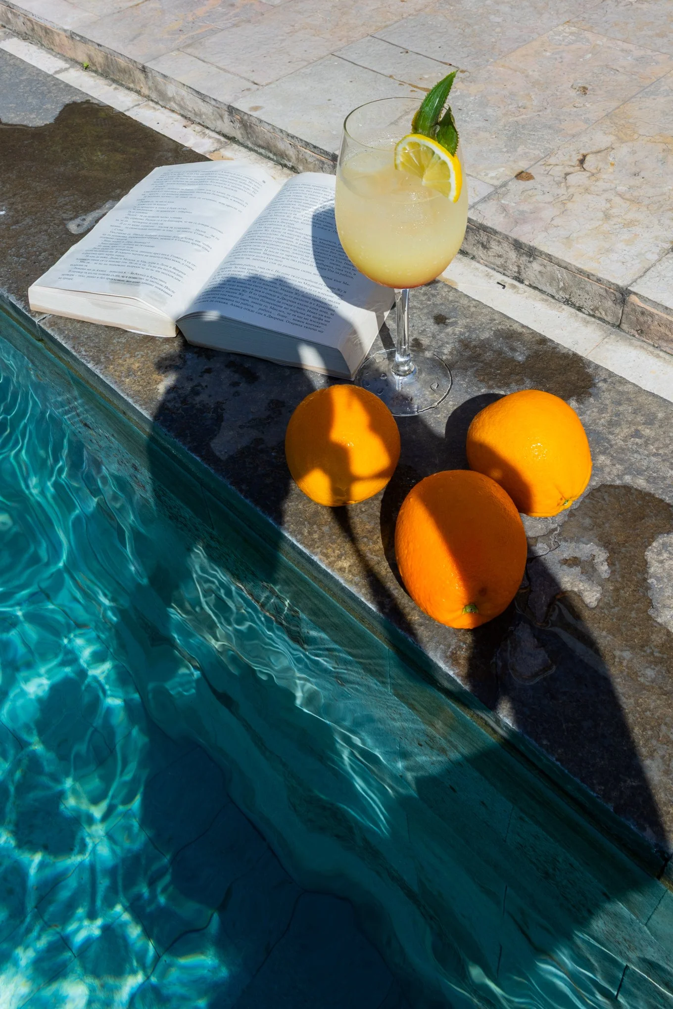A glass of lemonade with a lemon slice, a leaf, and a straw rests on the edge of a swimming pool. An open book lies next to the glass, and three oranges are placed nearby on the stone poolside. The pool water is blue, with sunlight creating ripples a