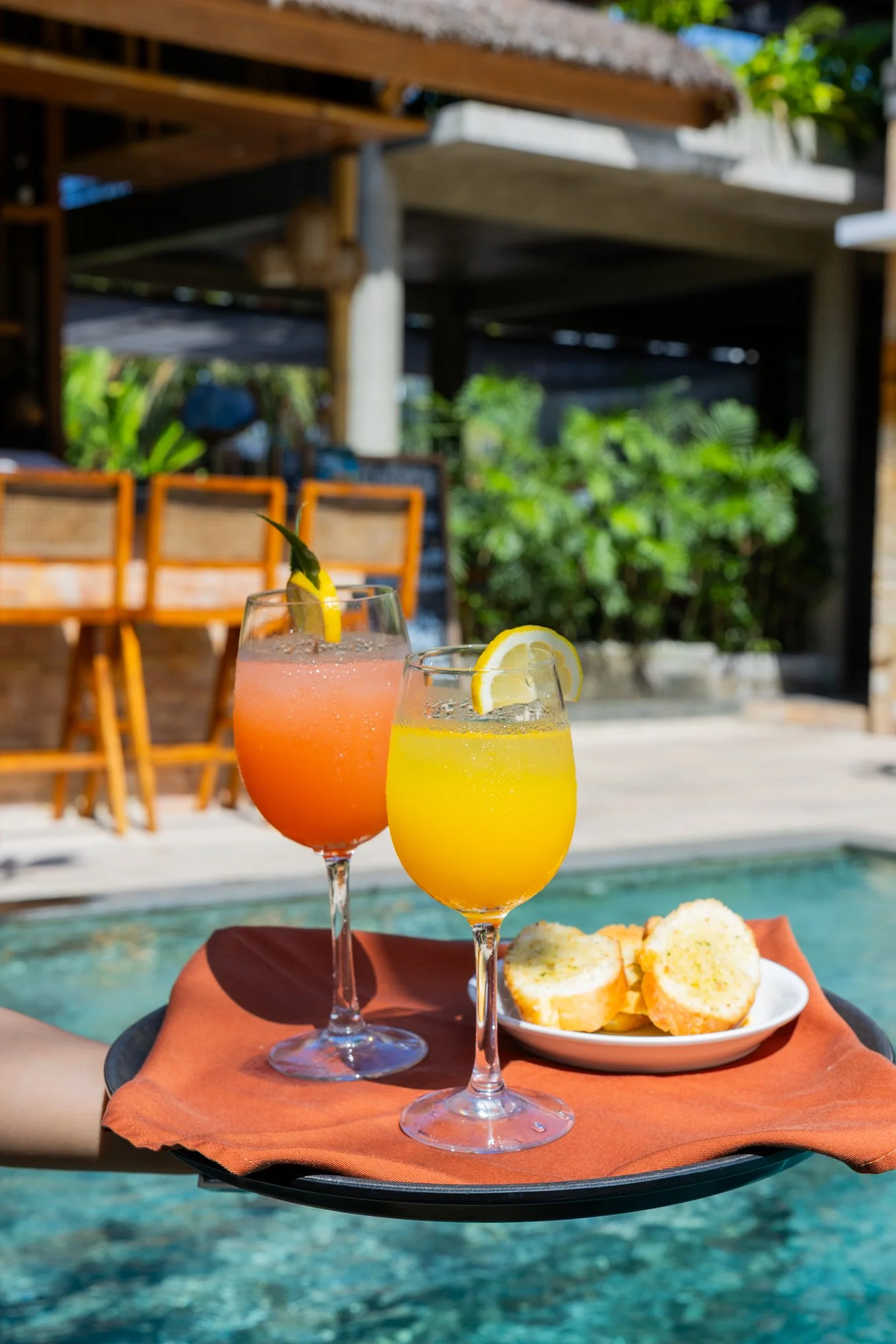 Two colorful cocktails with lemon slices on a floating tray, garnished with pineapple and mint, with bread rolls on a plate in the background, set outdoors near a pool.