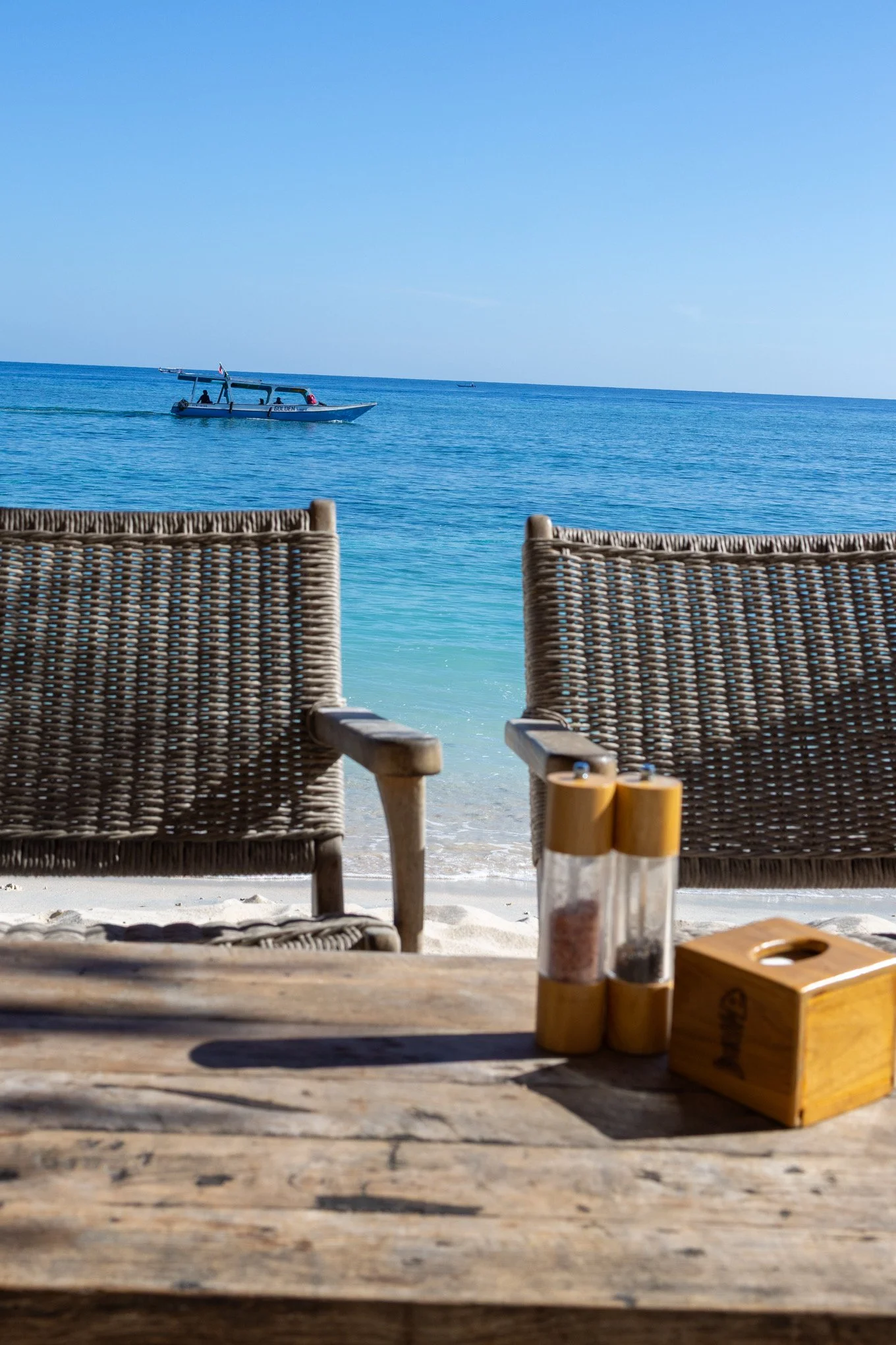 View of a beach with wicker chairs, a wooden table with spice jars, looking out to the ocean with a moving boat, clear blue sky, and calm water.