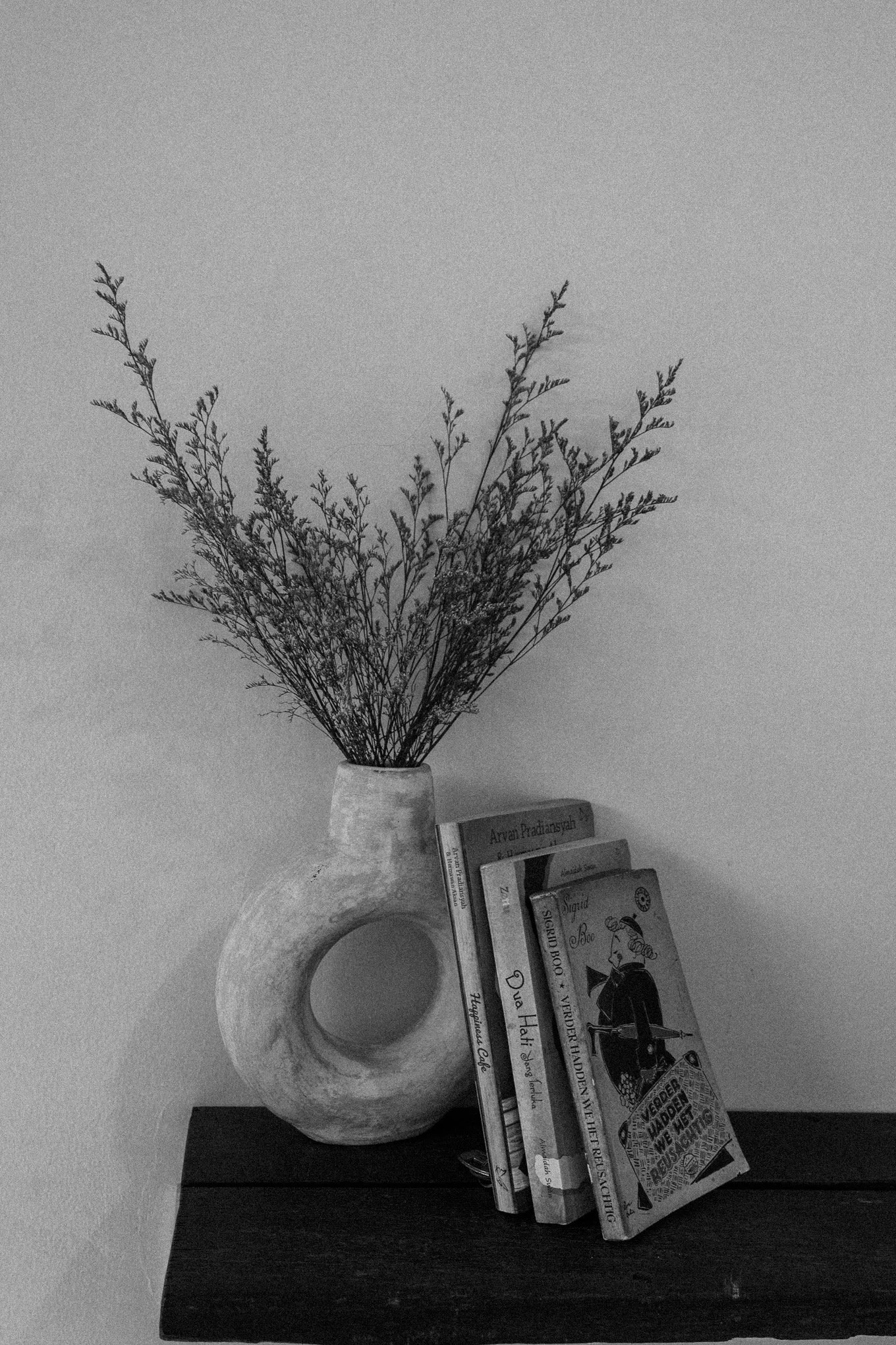 A ceramic vase with a circular opening at the base, holding dried branches. Beside the vase, there is a stack of books with visible titles, including 'Arvani Pradansyah' and others. A magazine featuring a woman in black attire on the cover leans agai