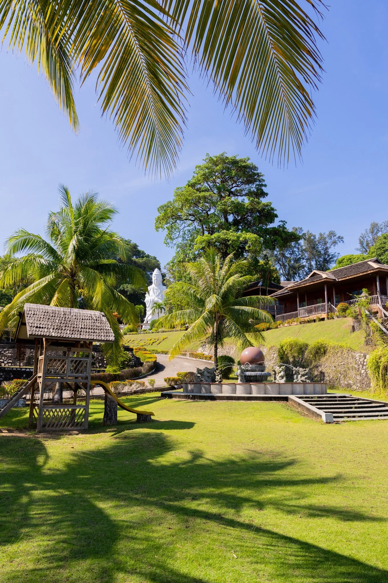 A garden with lush green grass, several palm trees, a white Buddha statue, and a building with a balcony, all under a clear blue sky.