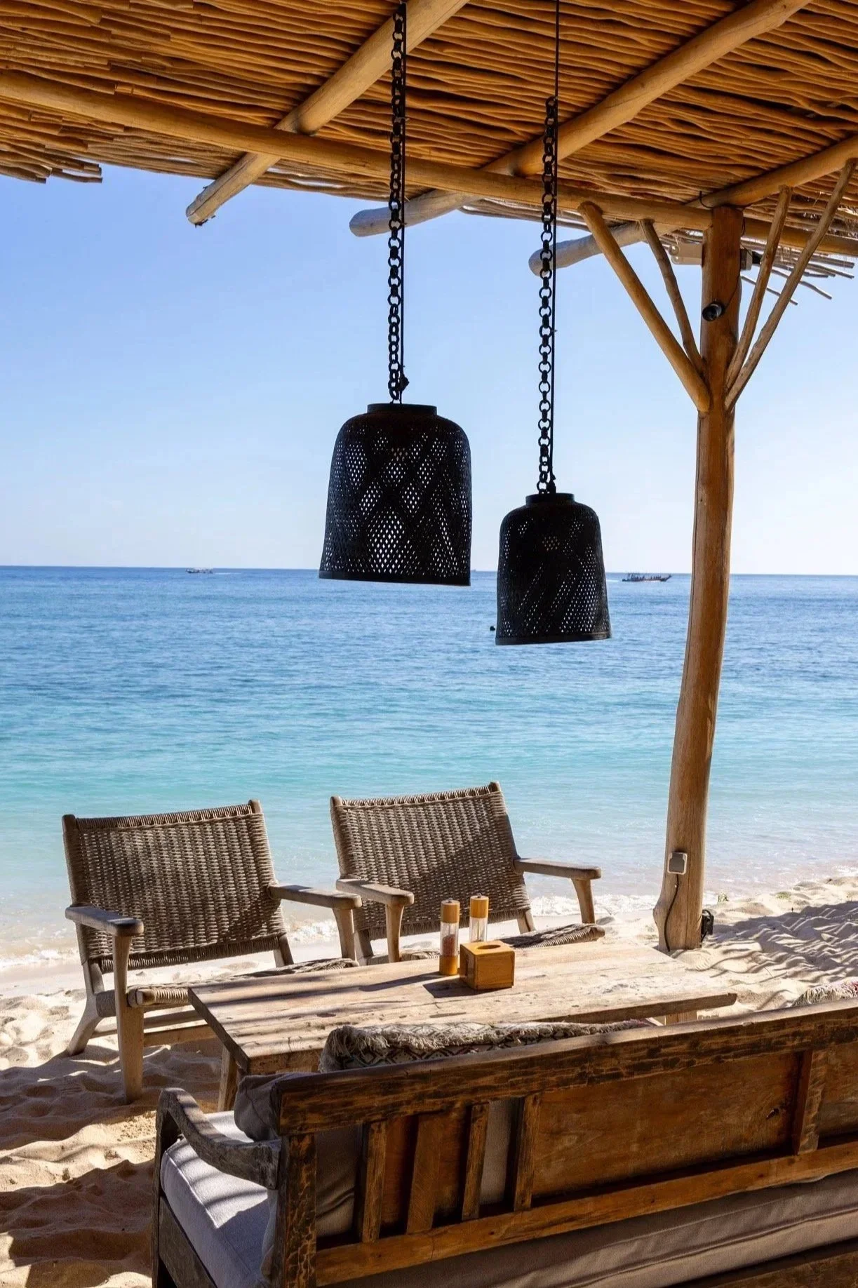Beach view with an outdoor seating area, two woven chairs, a wooden table with candles, under a thatched roof with hanging black lamps, overlooking clear blue ocean water and small boats in the distance.