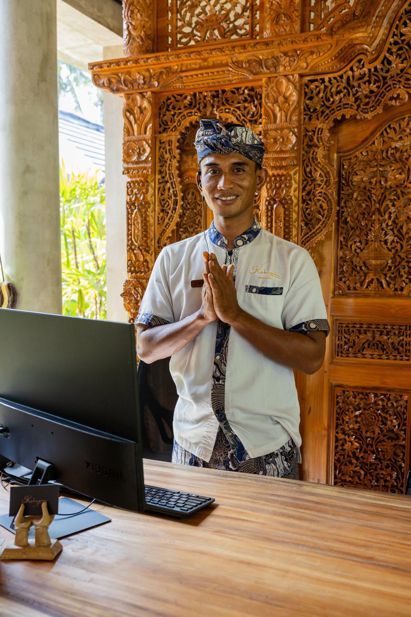 A smiling man wearing traditional Balinese attire and a headwrap, standing behind a desk with a computer, in front of a carved wooden backdrop.