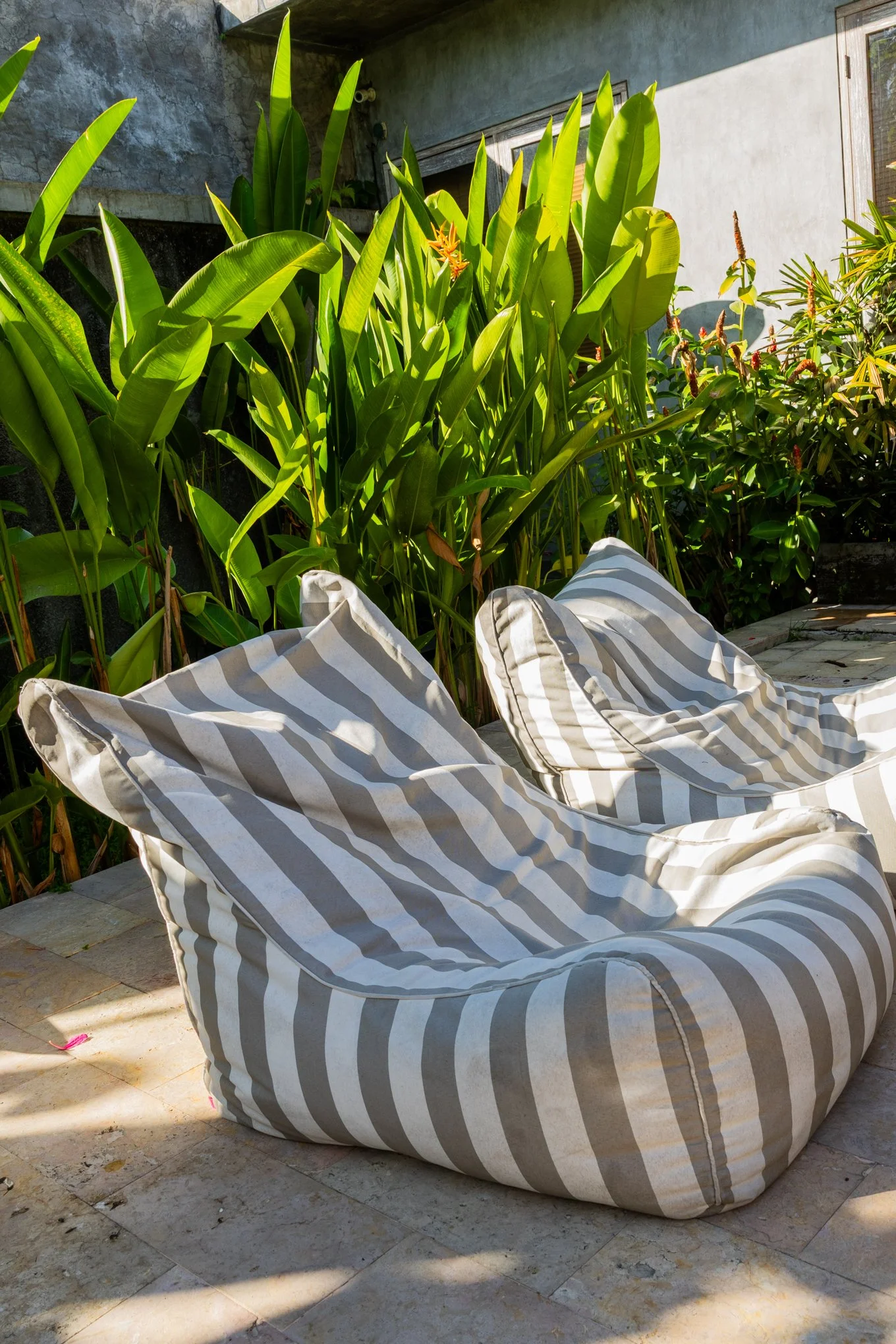 Sunlit outdoor patio with large striped bean bags and green tropical plants along a wall.