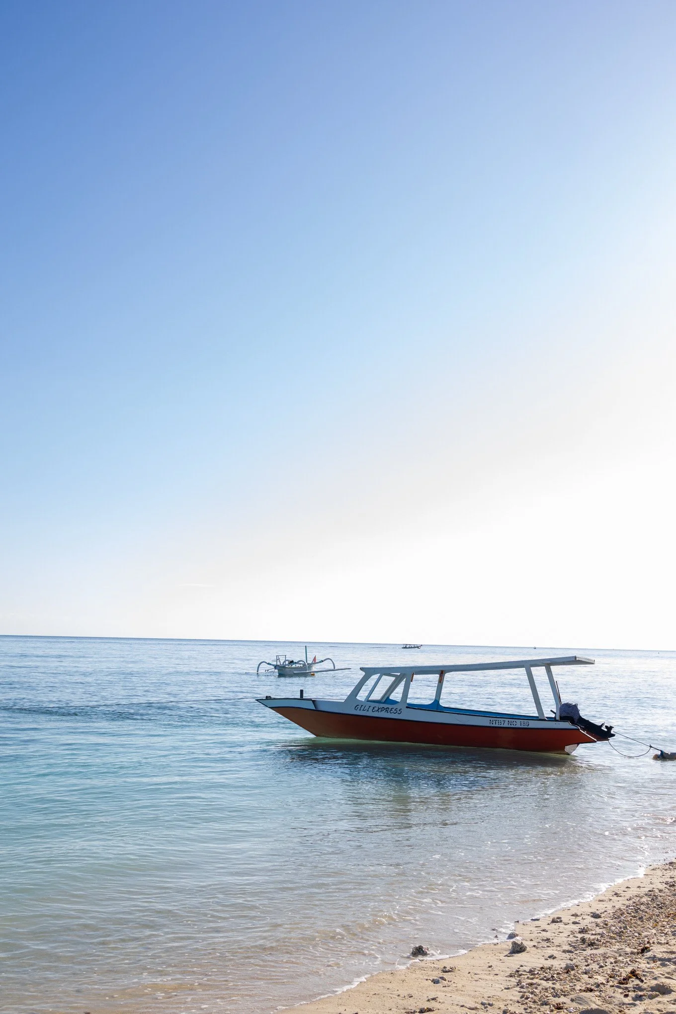 A beach scene with a small boat in the water, another boat in the distance, and a sandy shoreline with rocks, under a clear blue sky.