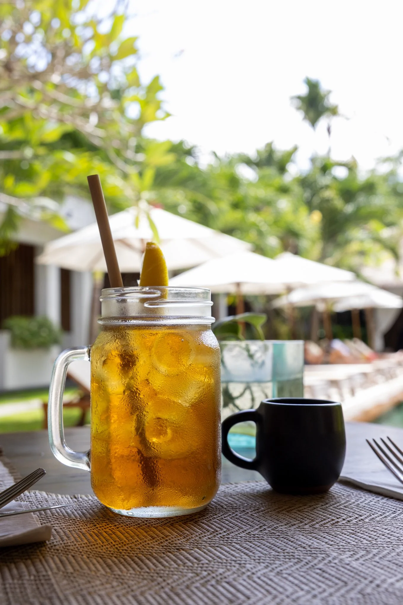 A glass jar filled with ice and iced tea, garnished with a lemon wedge and a straw, on a woven tablecloth at an outdoor setting with umbrellas and greenery in the background.