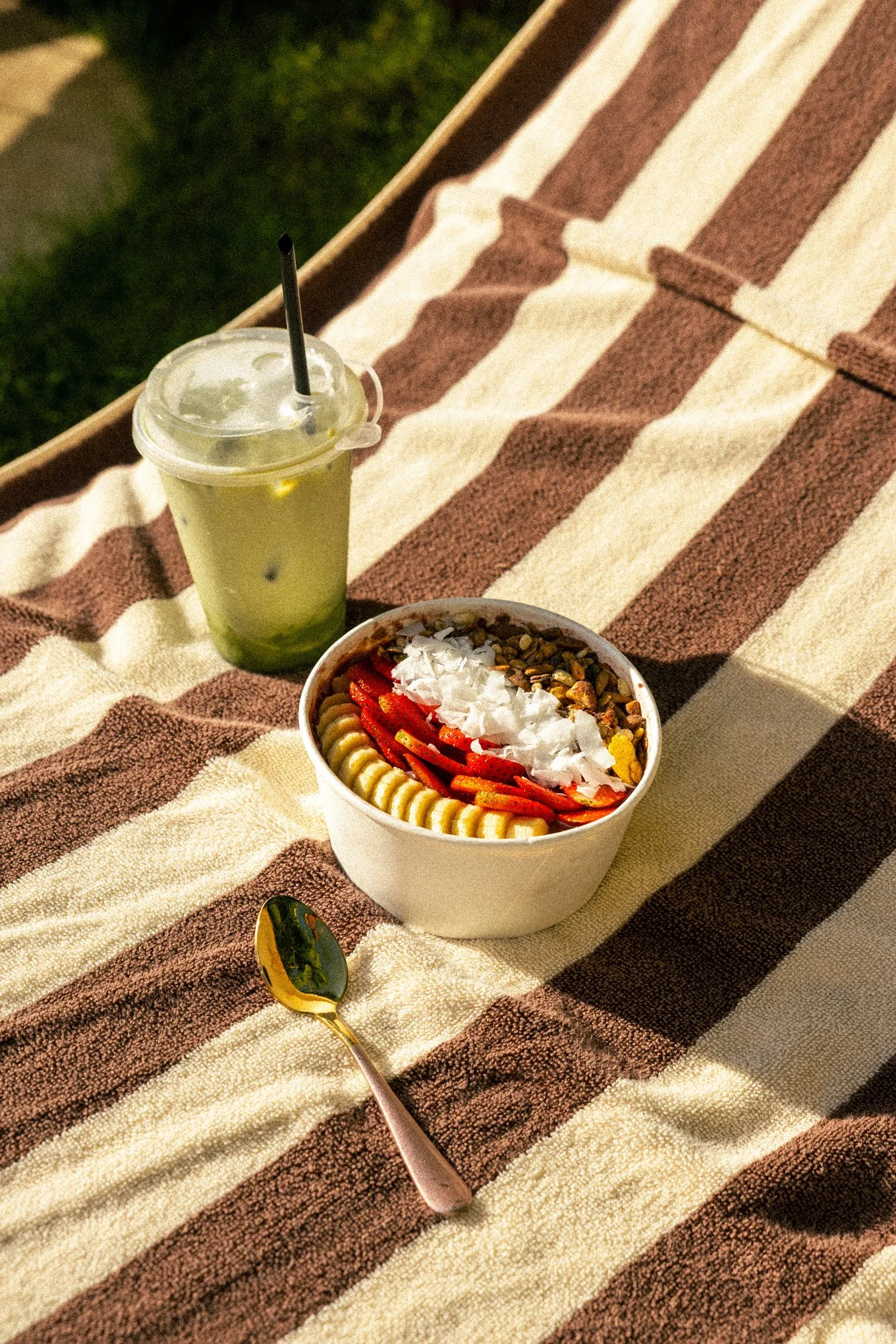A striped brown and white towel on grass, with a bowl of sliced strawberries, banana, shredded coconut, and granola, a gold spoon, and a cold drink with ice and a black straw.