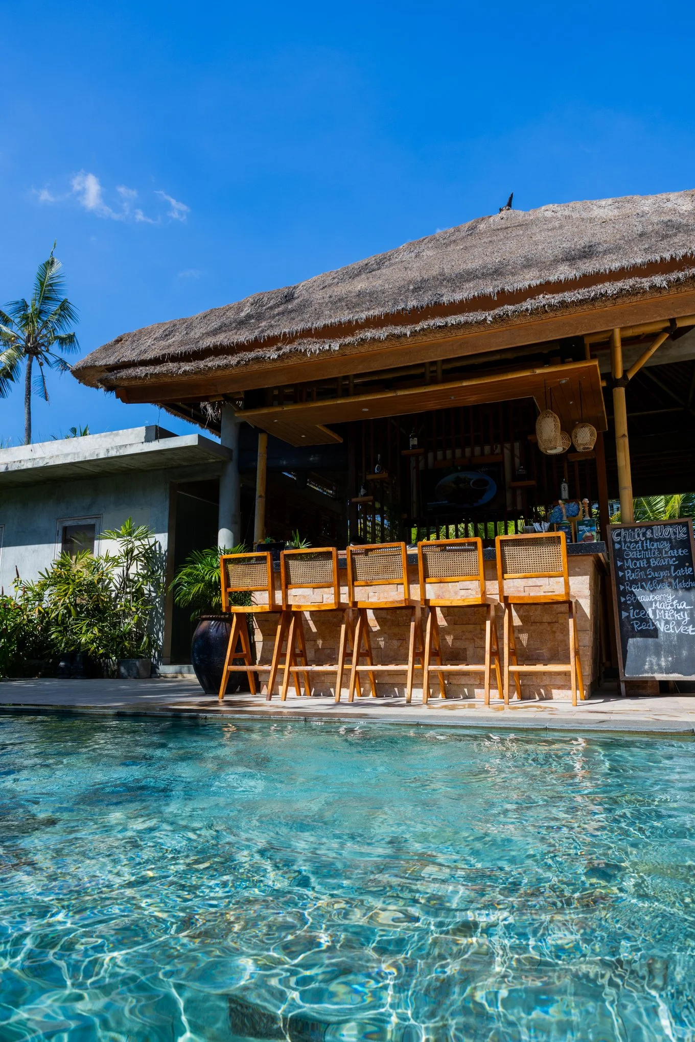 An outdoor pool area of a tropical resort with tall palm trees, thatched roof hut, and a clear blue sky.