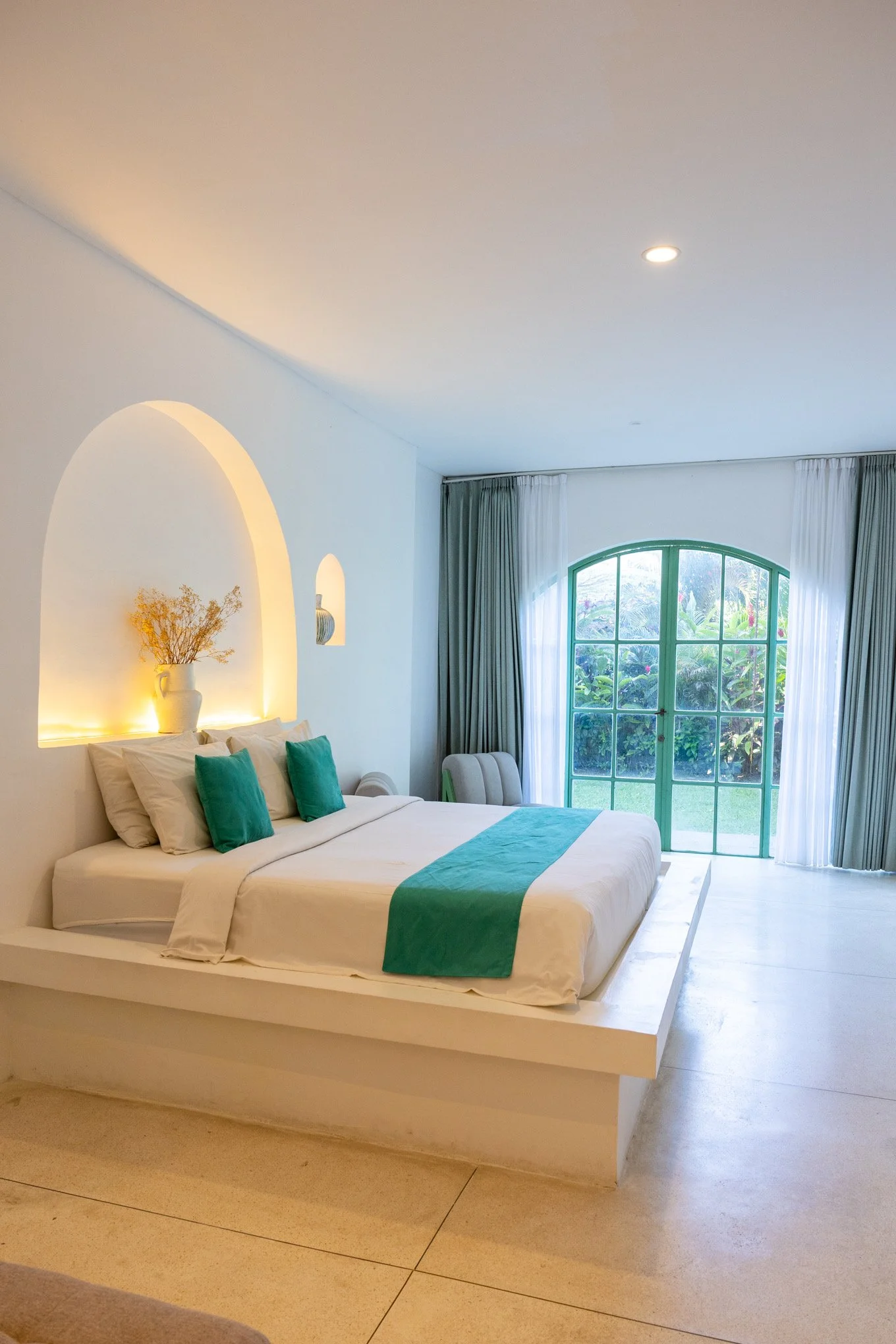 Bright and airy bedroom with a large white bed that has teal accent pillows and a teal runner. The room features an arched green-framed window with white curtains, a built-in wall niche with a vase and dried flowers, and soft natural light.
