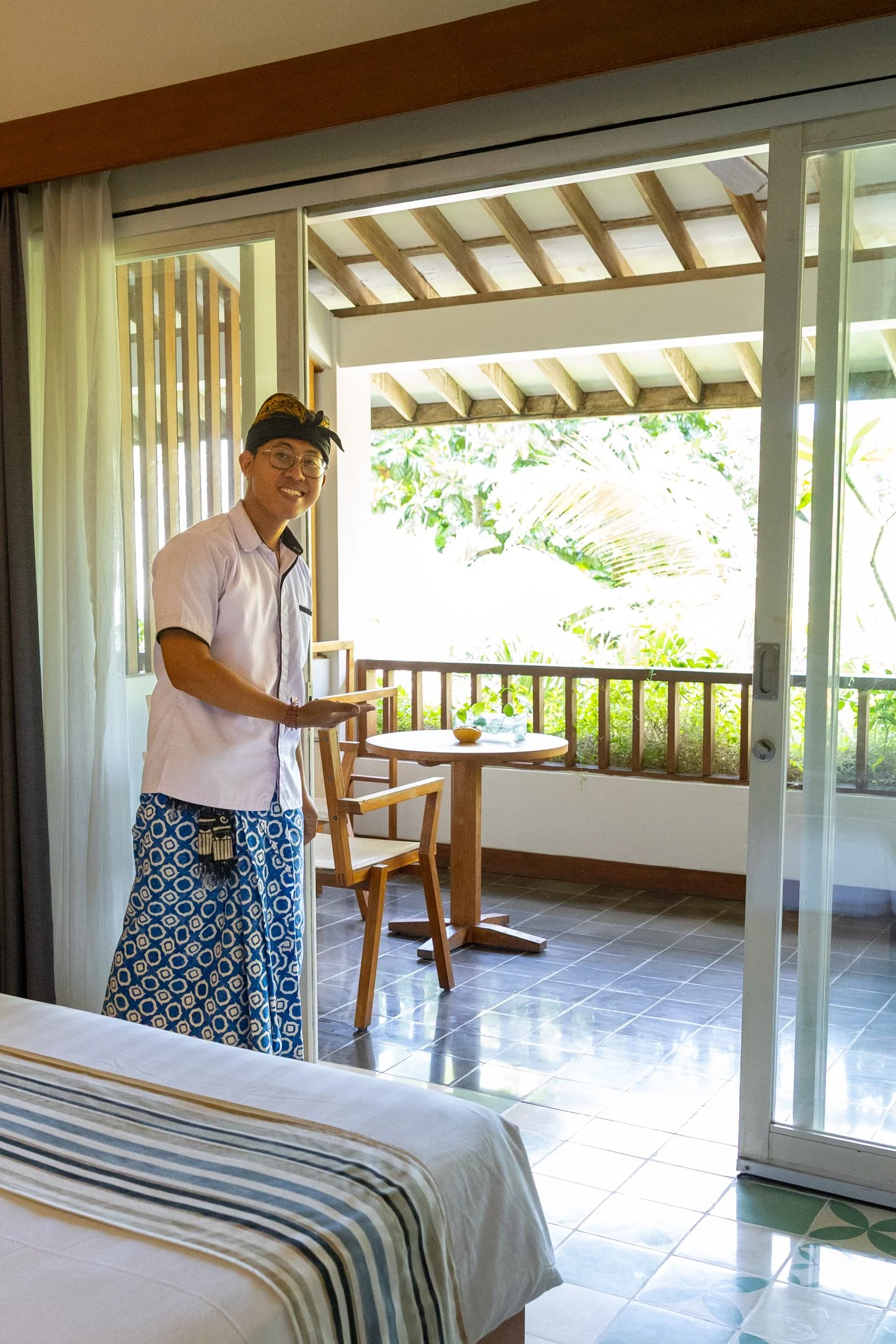 A man wearing a white shirt, patterned blue and white pants, glasses, and a traditional Indonesian headwrap is standing inside a room, smiling at the camera. The room has a balcony with wooden beams and green foliage outside. There is a round table w