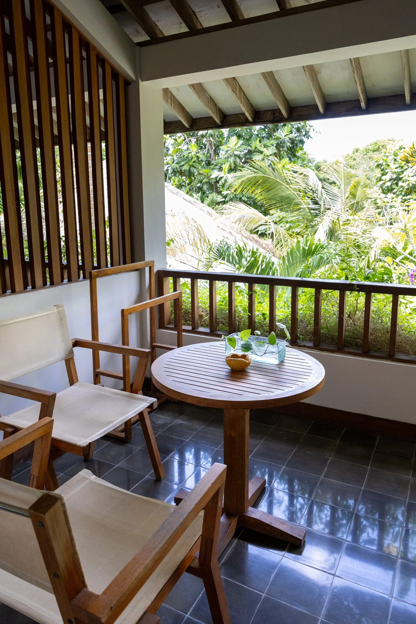 Balcony with wooden chairs and round table, overlooking lush green tropical plants and trees.