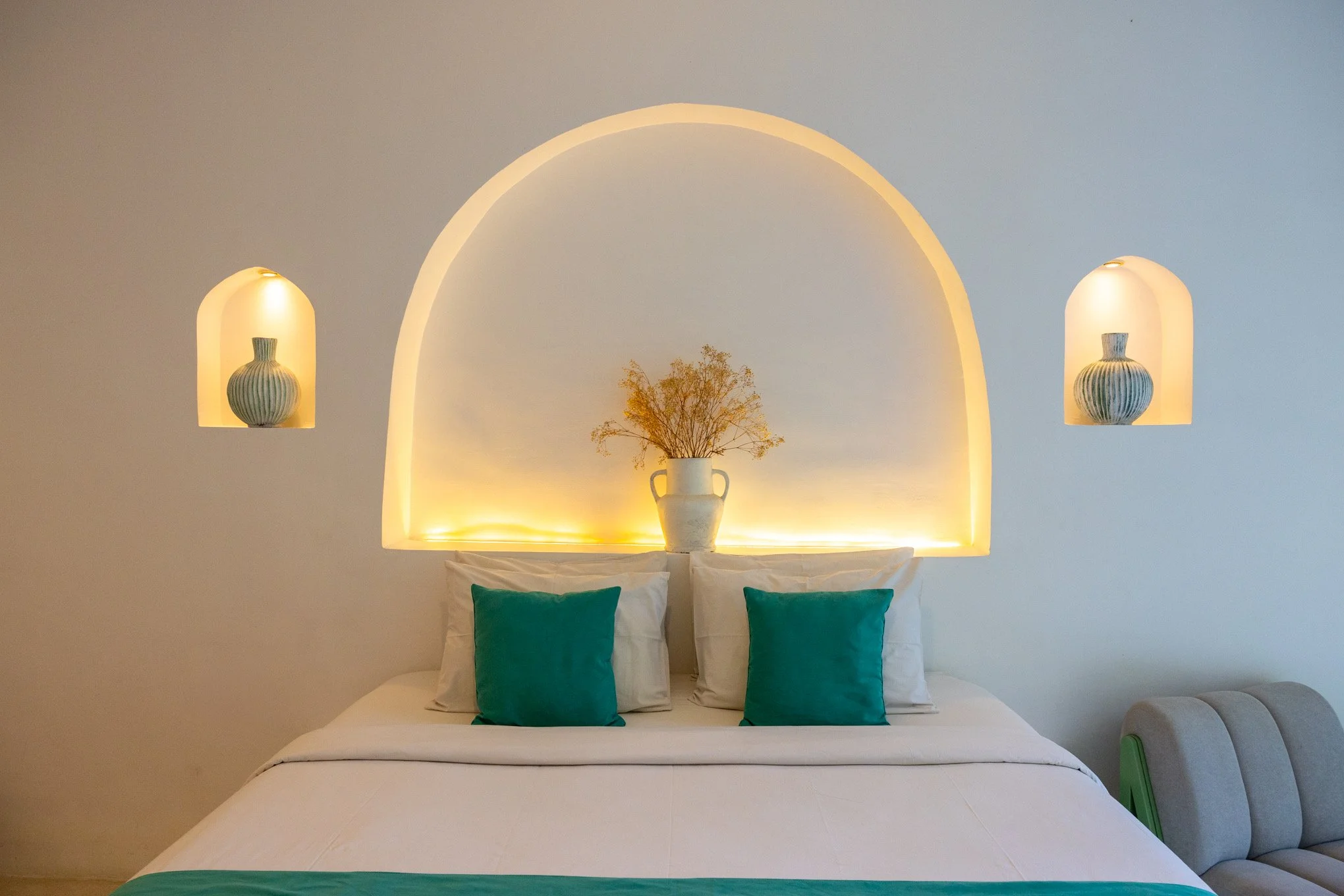 A neatly made bed with white linens and four teal-colored pillows, set against a light-colored wall with built-in wall niches. The central niche contains a white vase with dried flowers, flanked by two smaller niches each holding a decorative vase. T