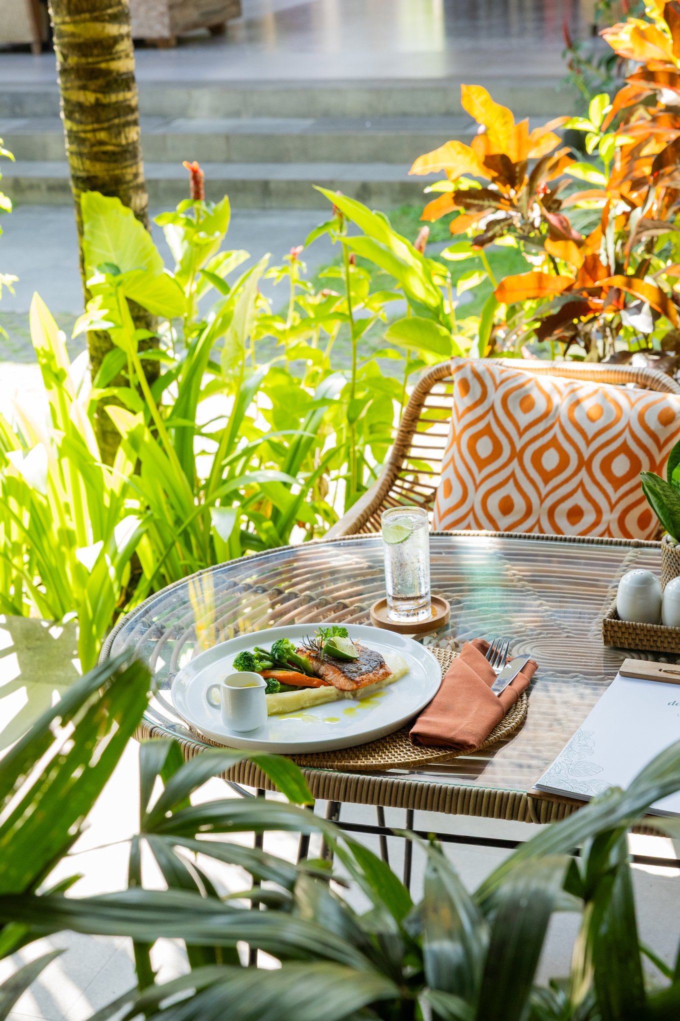 A table with a plated meal featuring cooked fish, vegetables, and sauce in an outdoor setting surrounded by green tropical plants and orange cushions on a chair, with steps and a pathway in the background.