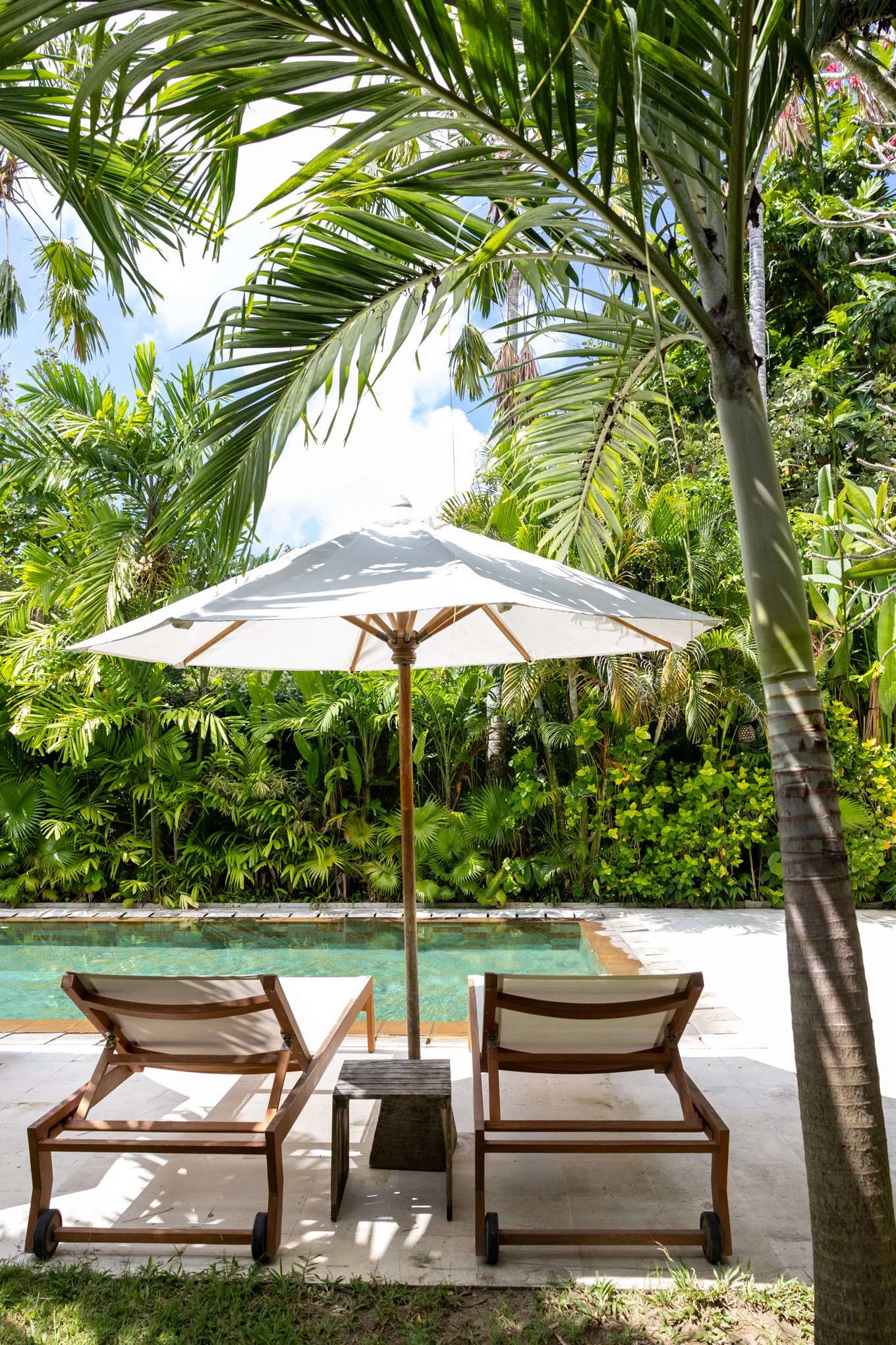 Poolside with two wooden lounge chairs, a white umbrella, surrounded by lush tropical green foliage, including palm trees, near a swimming pool.