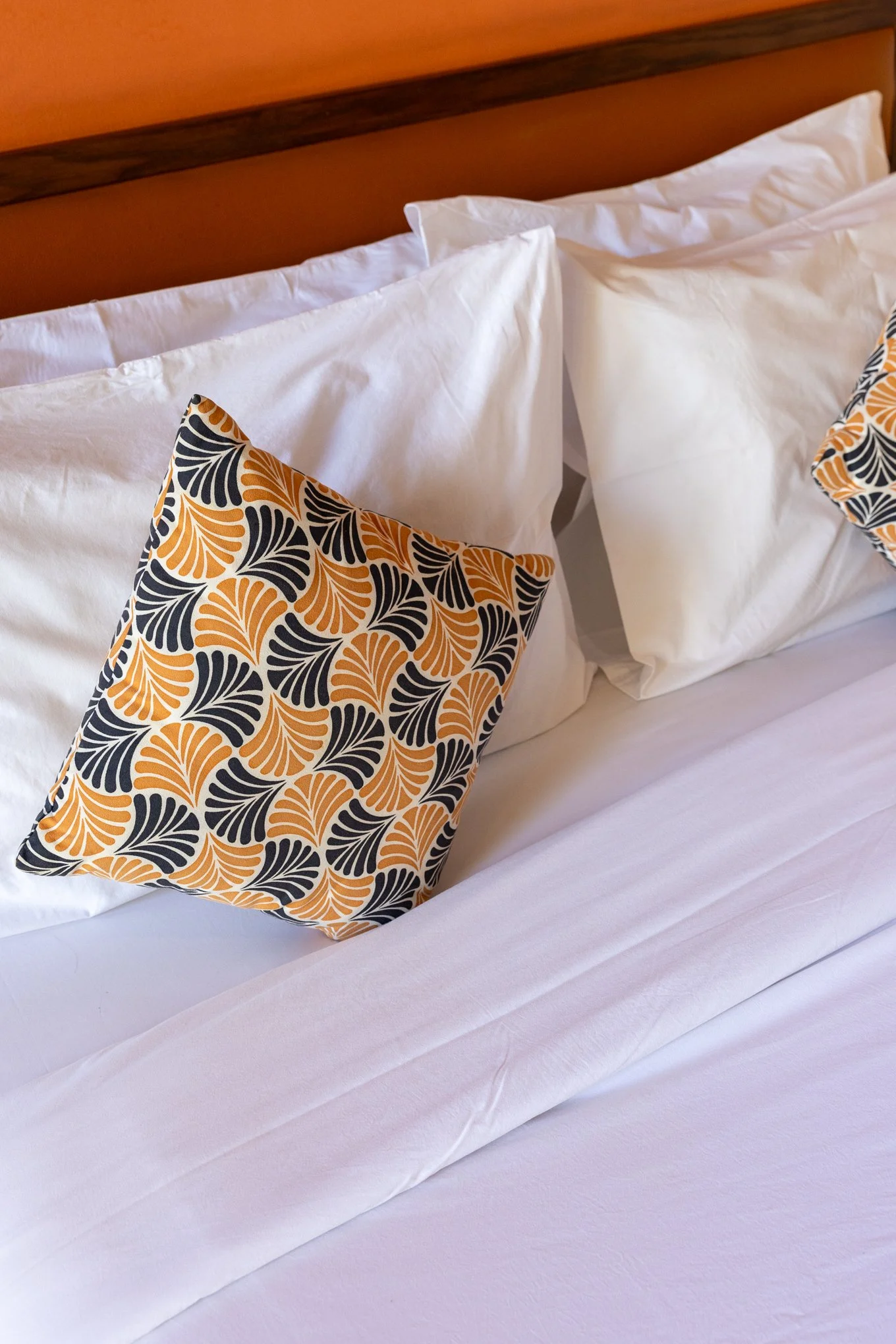 Close-up of a bed with white sheets, white pillows, and decorative pillows with an orange, black, and white pattern. A wooden headboard is visible in the background.