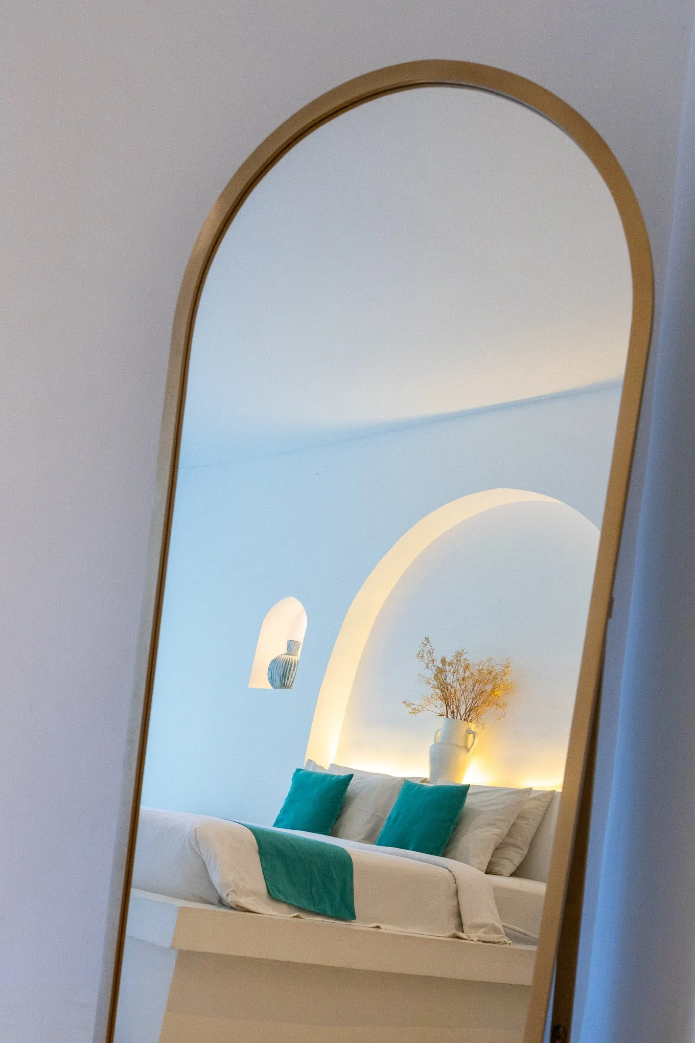 A bedroom with a white bed, a white vase with a tree and flowers, framed via a mirror with a gold border, with a blue wall and a ceiling light.