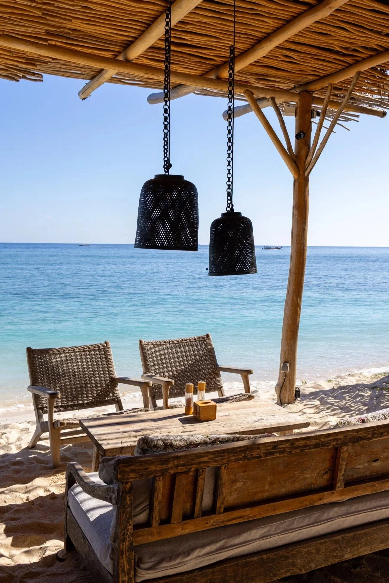 Beachside seating area with wicker chairs and wooden table, overlooking the ocean under a thatched roof. Two black lanterns hang from the roof, and the sky is clear with a few boats visible in the distance.