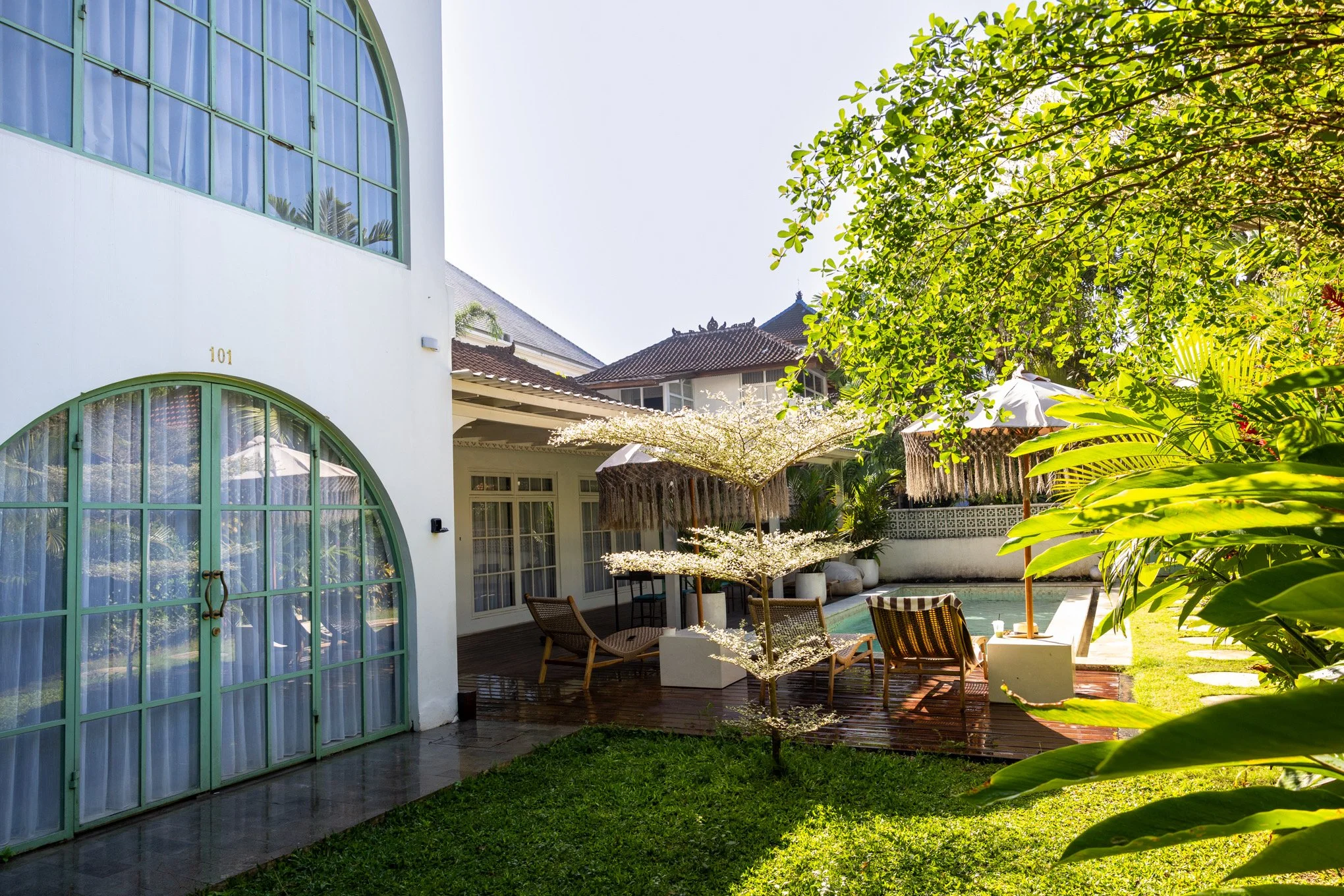 A backyard scene featuring a modern white house with large green-framed arched windows, lush green trees, and outdoor furniture under umbrellas, with a swimming pool and a garden area.
