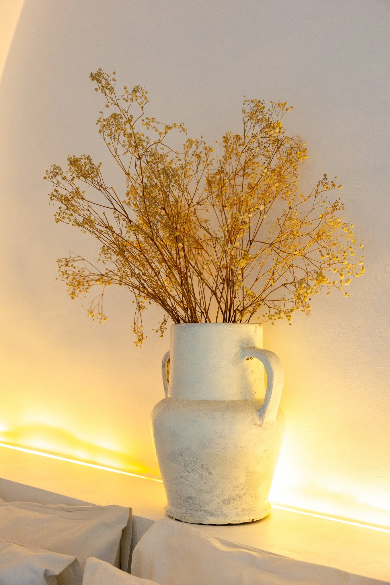 A white ceramic vase with a handle, filled with dried baby's breath flowers, placed on a surface against a wall with soft warm lighting.