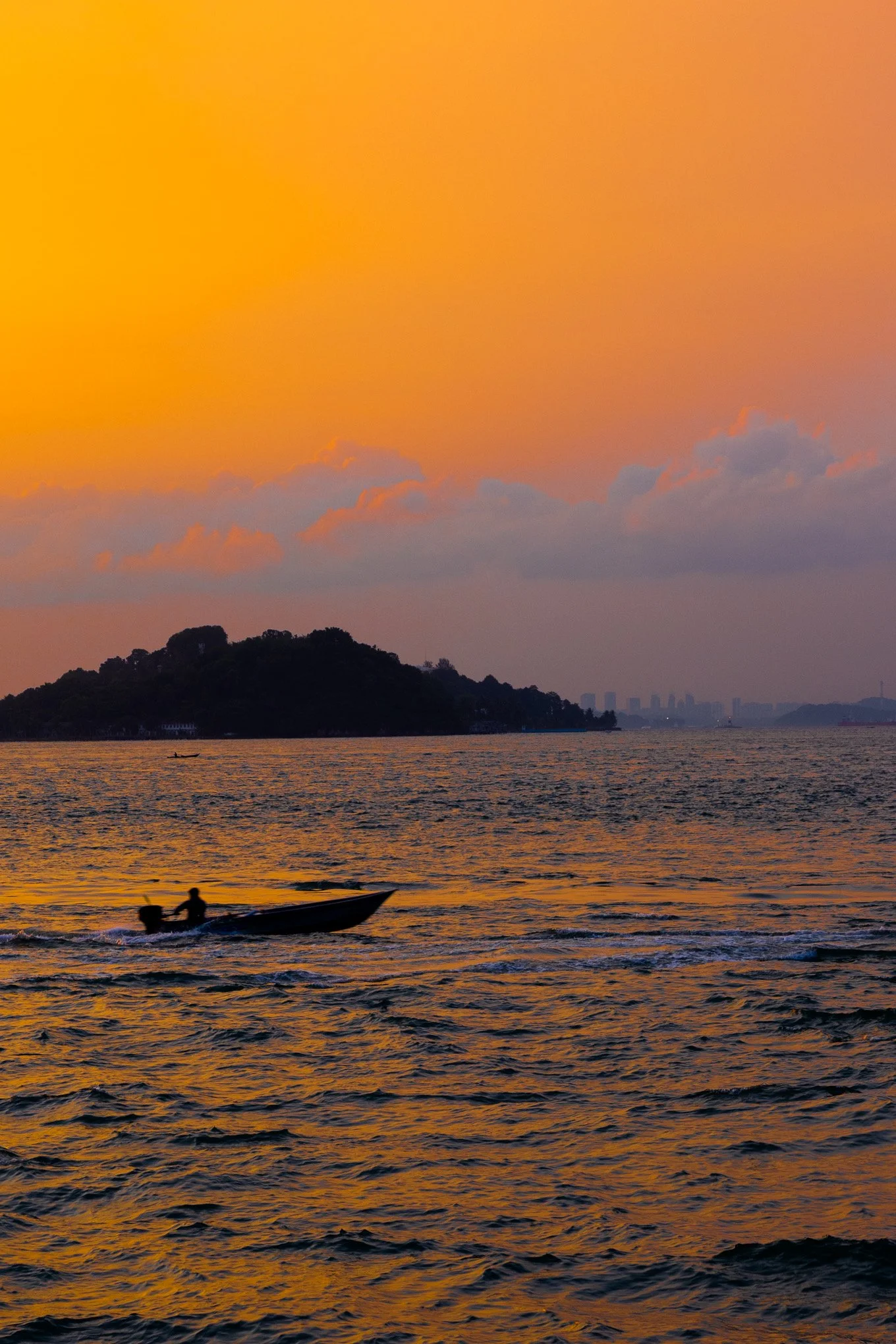 Sunset over a body of water with a small boat and two people in the foreground, a hilly landmass with trees, and buildings in the distance under a colorful sky with clouds.